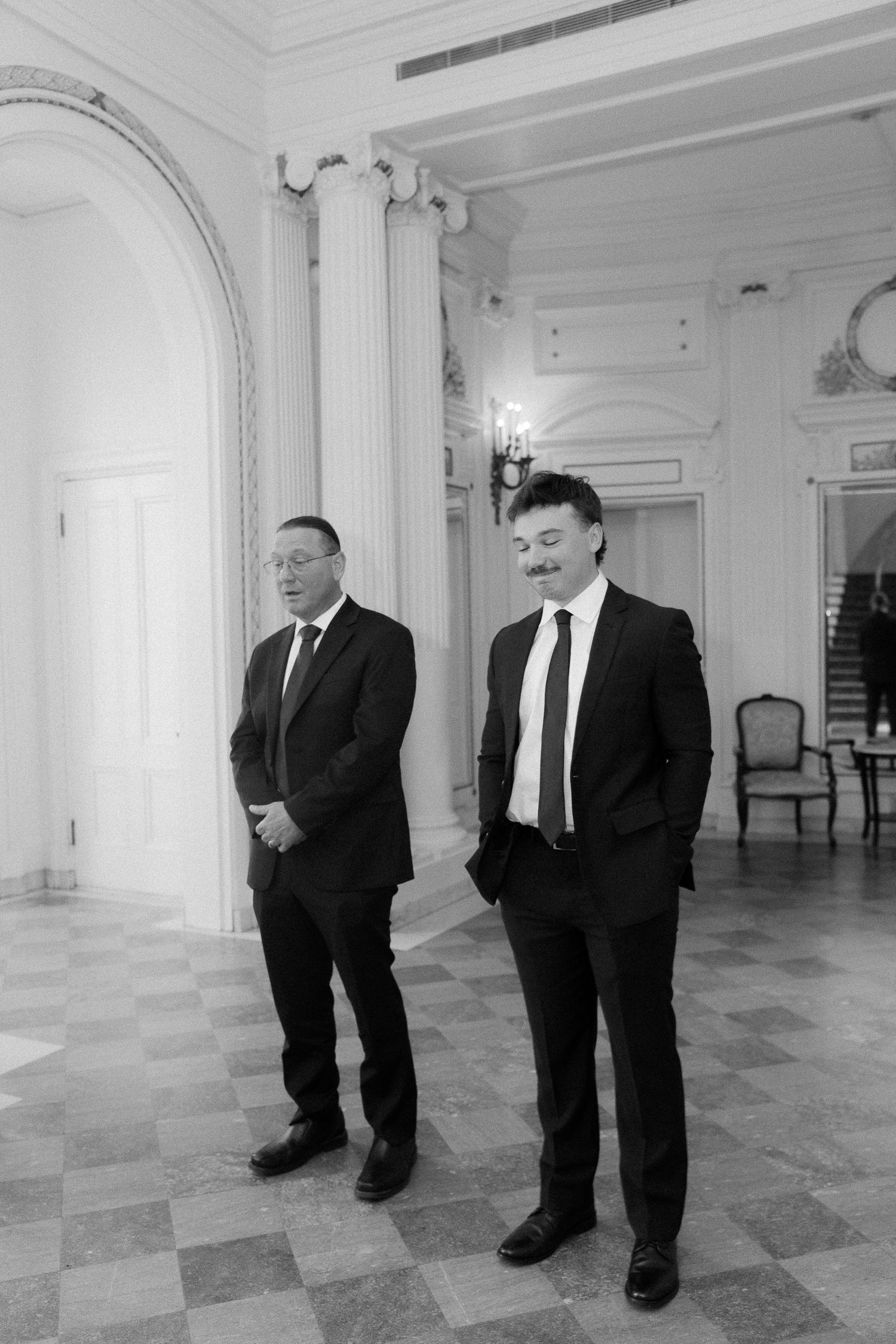 Two men in black suits stand in elegant white ballroom with tall columns at a formal event.