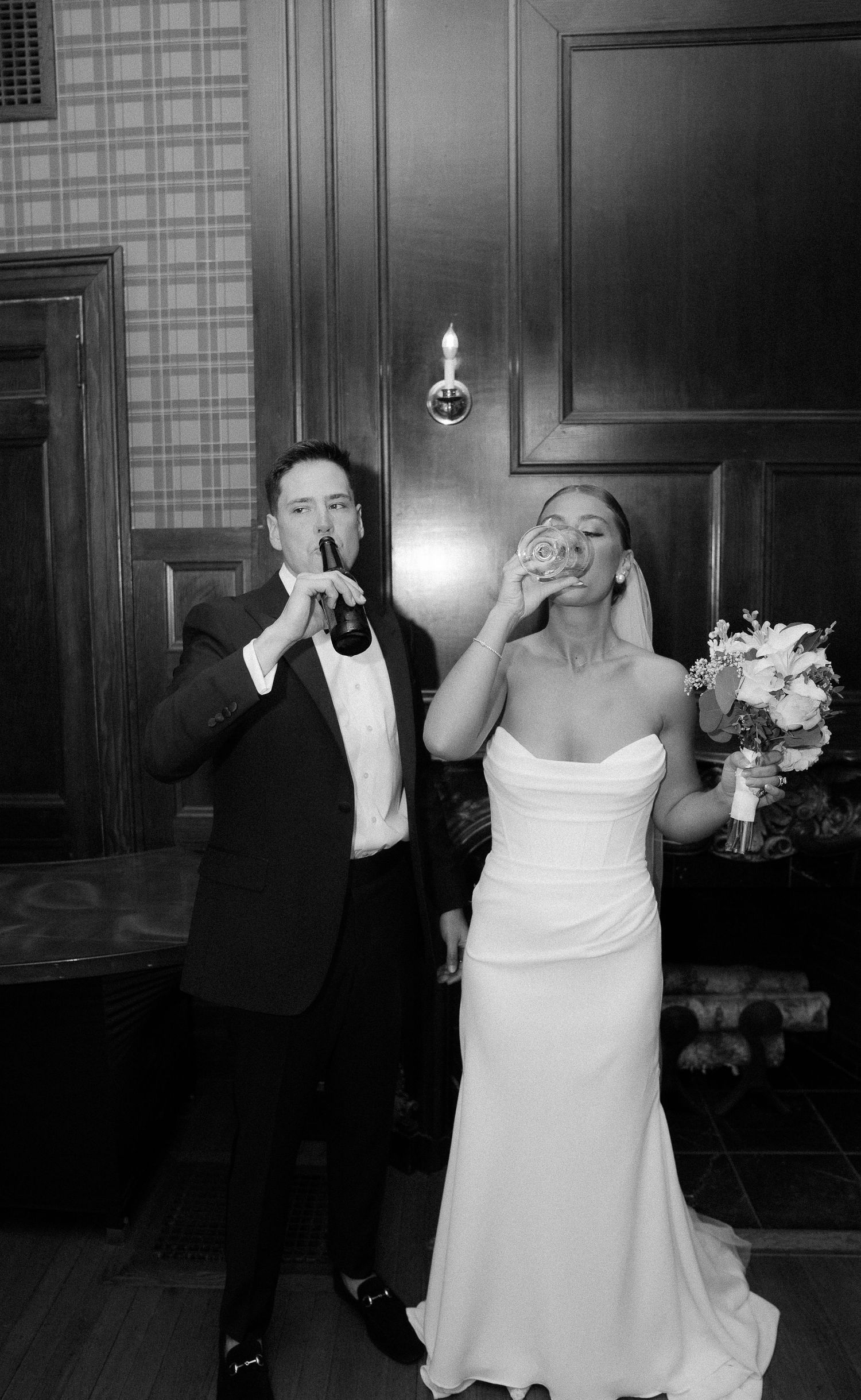 Black and white photo of newlyweds celebrating with drinks, groom sipping beer and bride raising a glass.