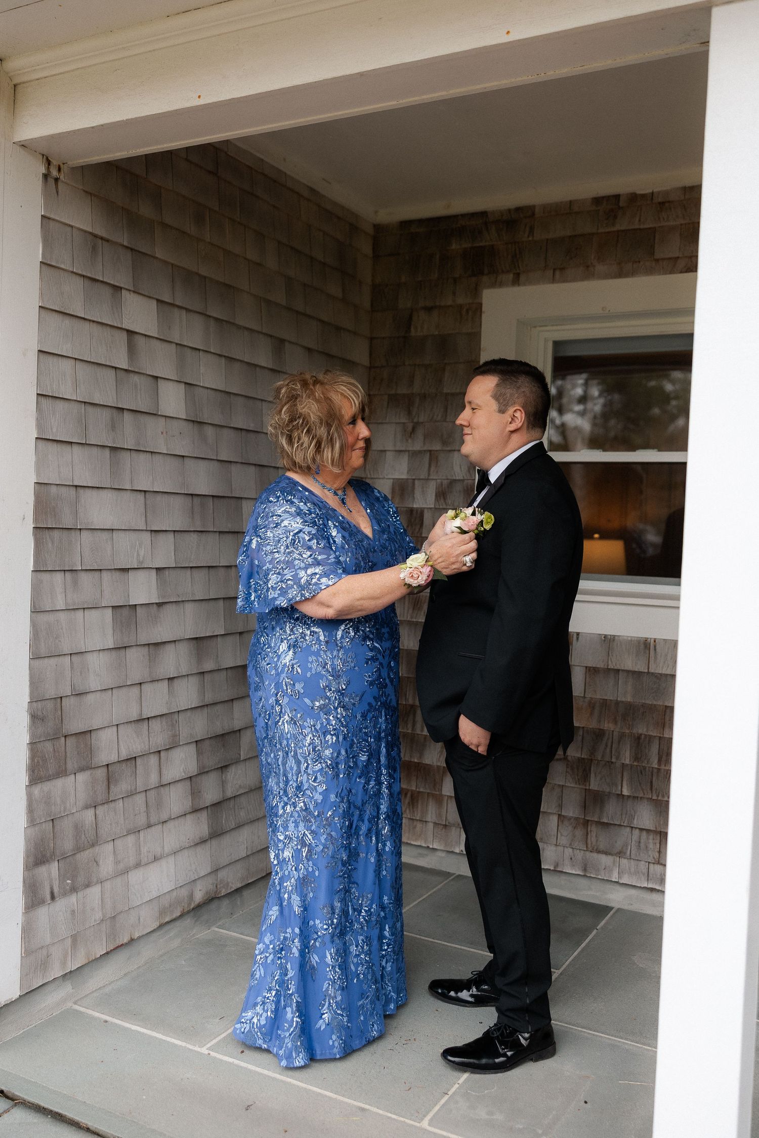 Woman in blue lace gown pins boutonniere on groom in black tuxedo outside a shingle-style building.