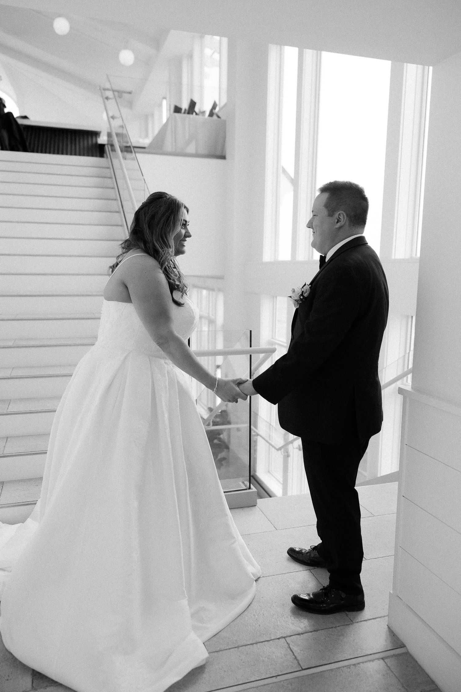 Black and white photo of a bride and groom holding hands on a staircase during their wedding first look.