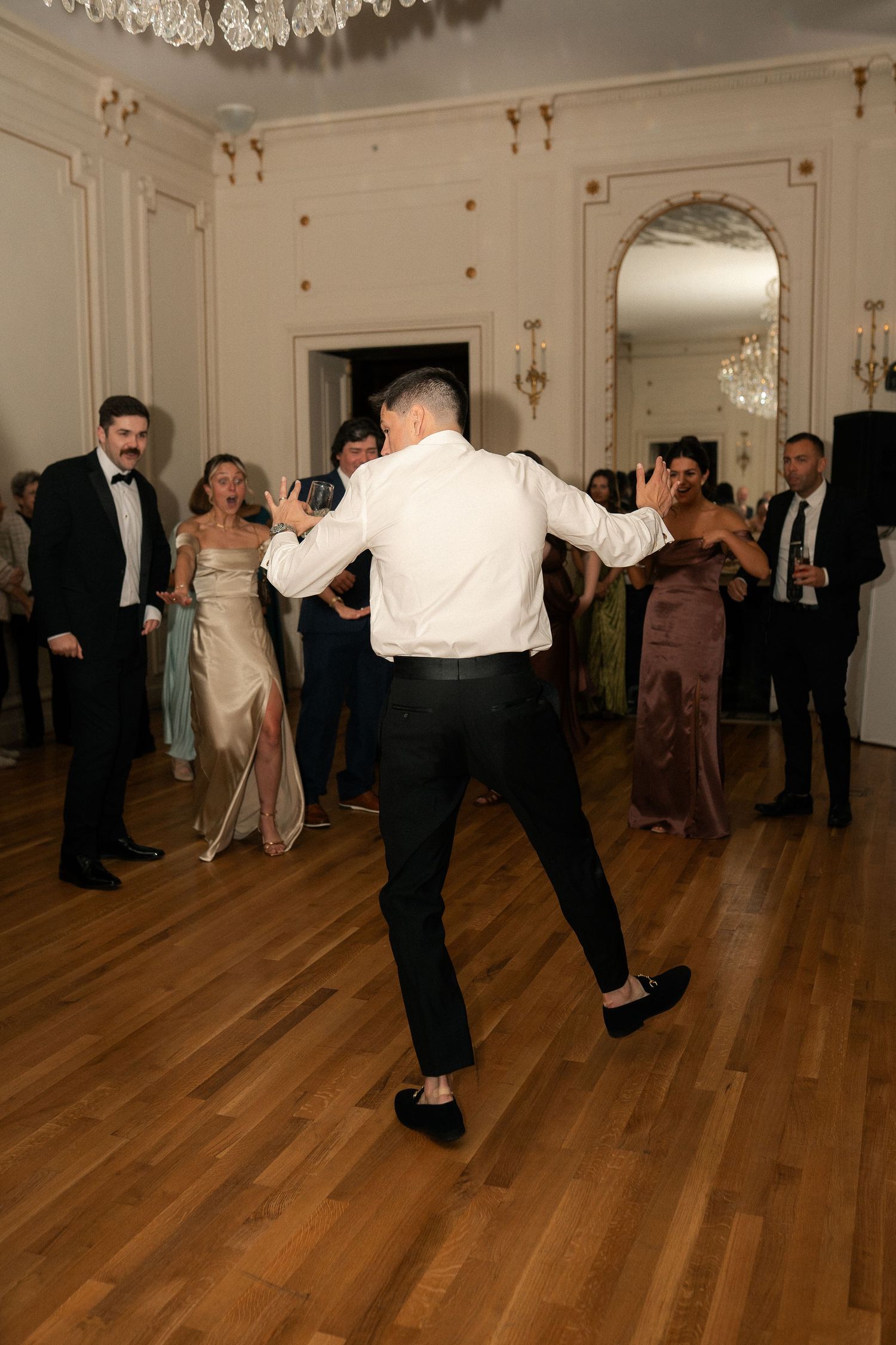Guests dancing at an elegant wedding reception in a grand ballroom with chandeliers and hardwood floors.