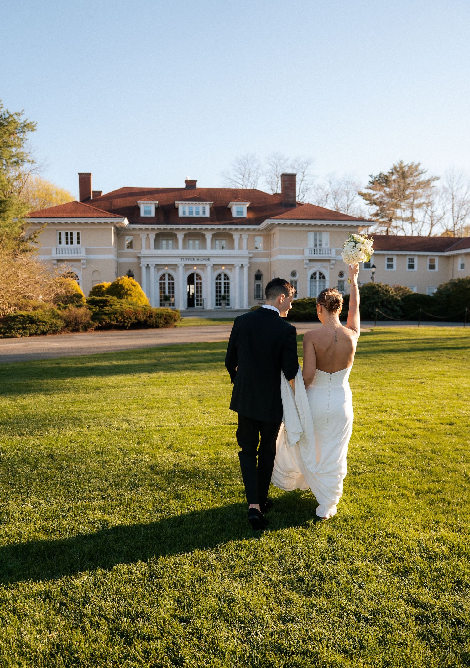 Bride and groom walk toward elegant mansion venue on lush green lawn on their wedding day.