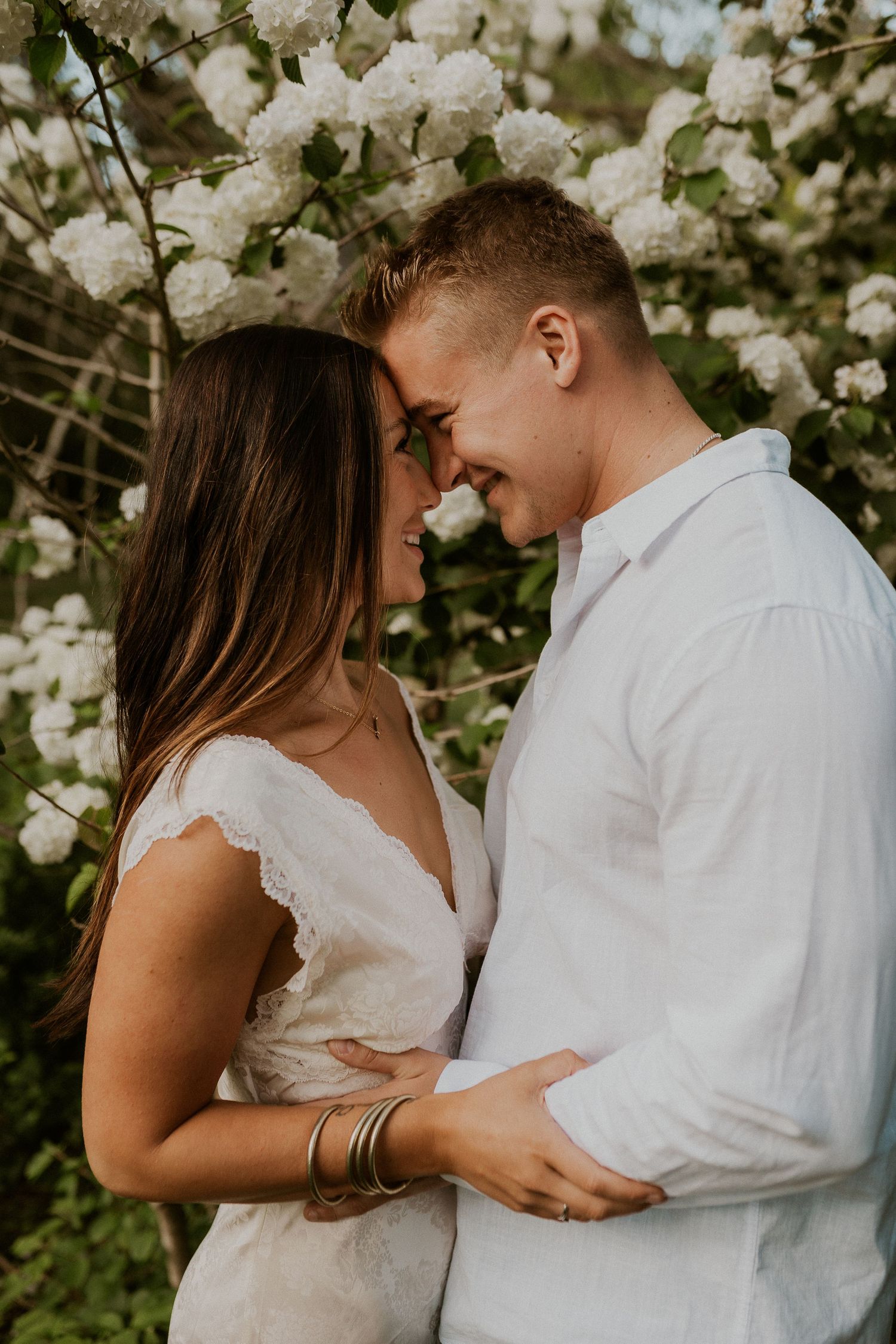 Romantic couple shares tender moments among blooming white flowers in a garden setting.