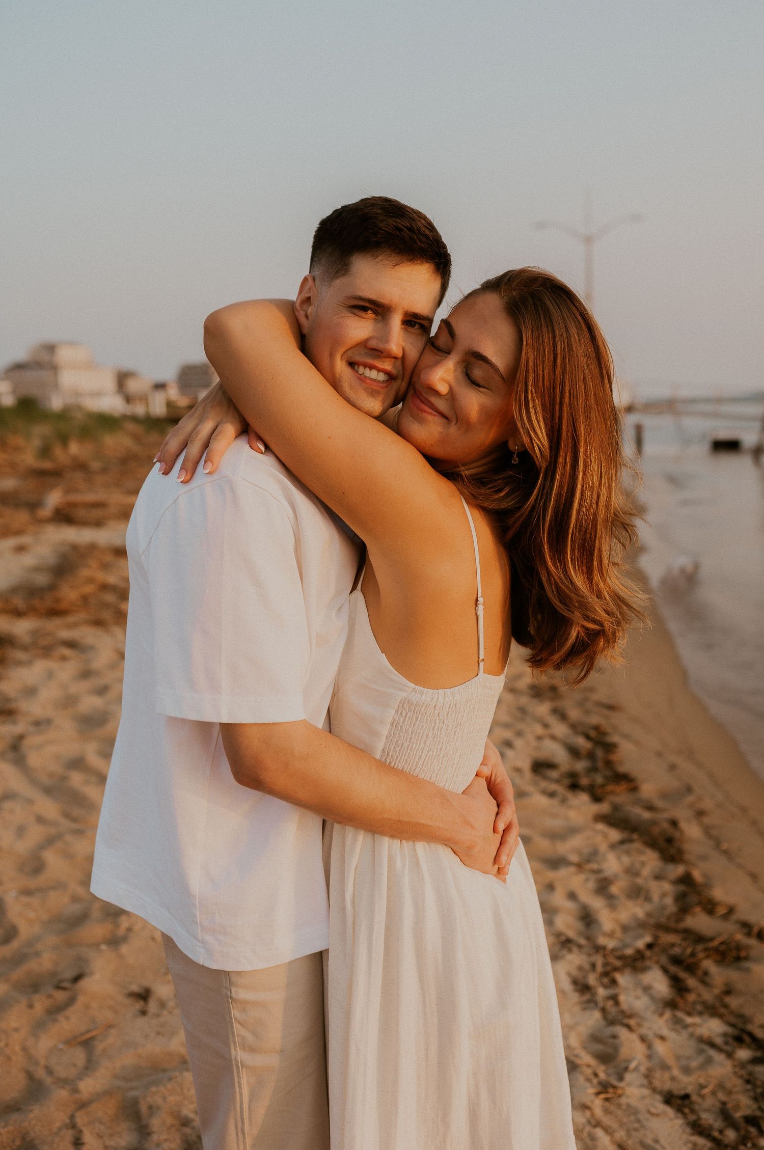 Warm-toned beach portrait of couple laughing and embracing in white attire.