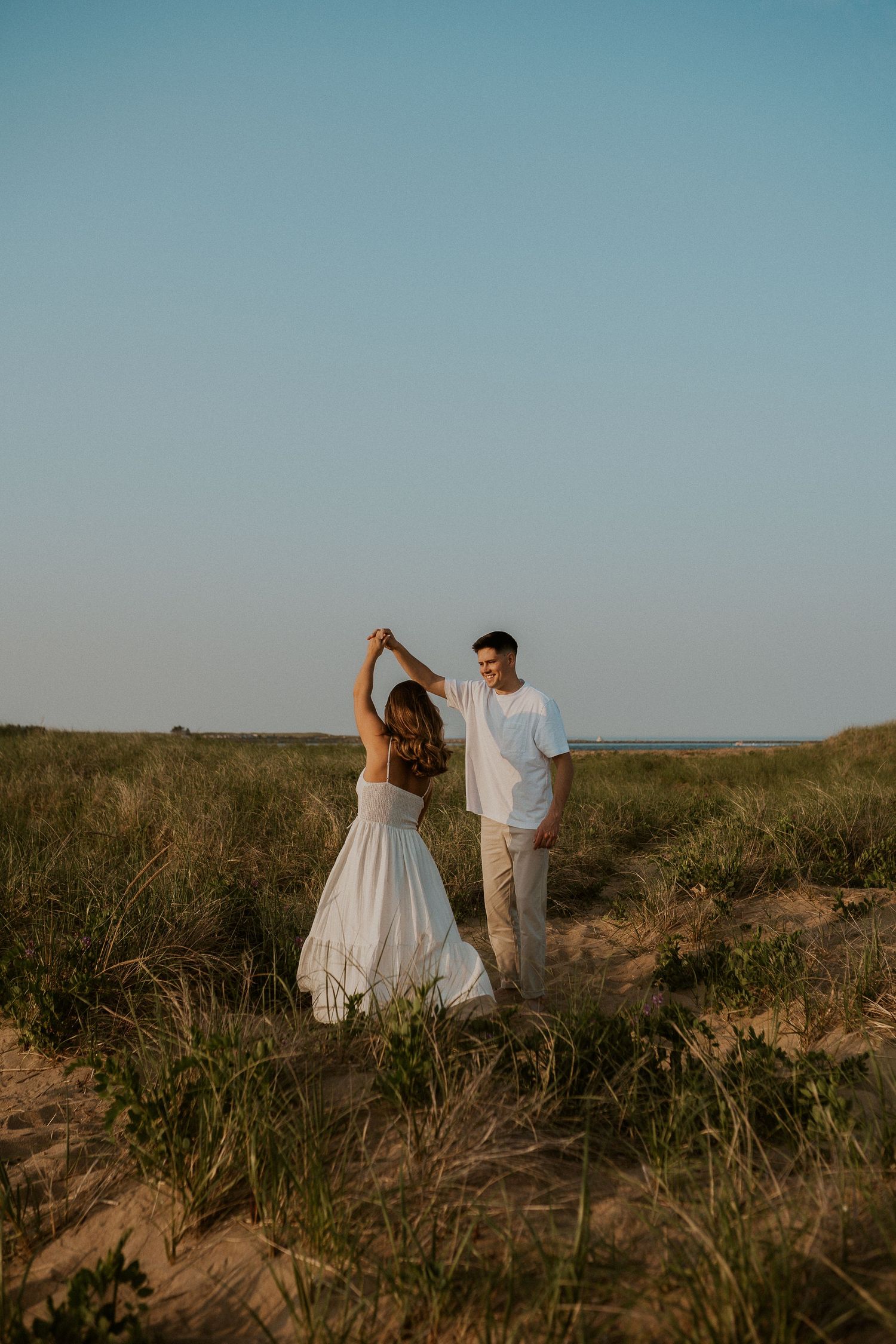 Couple in white clothing dancing on coastal dunes against blue sky.