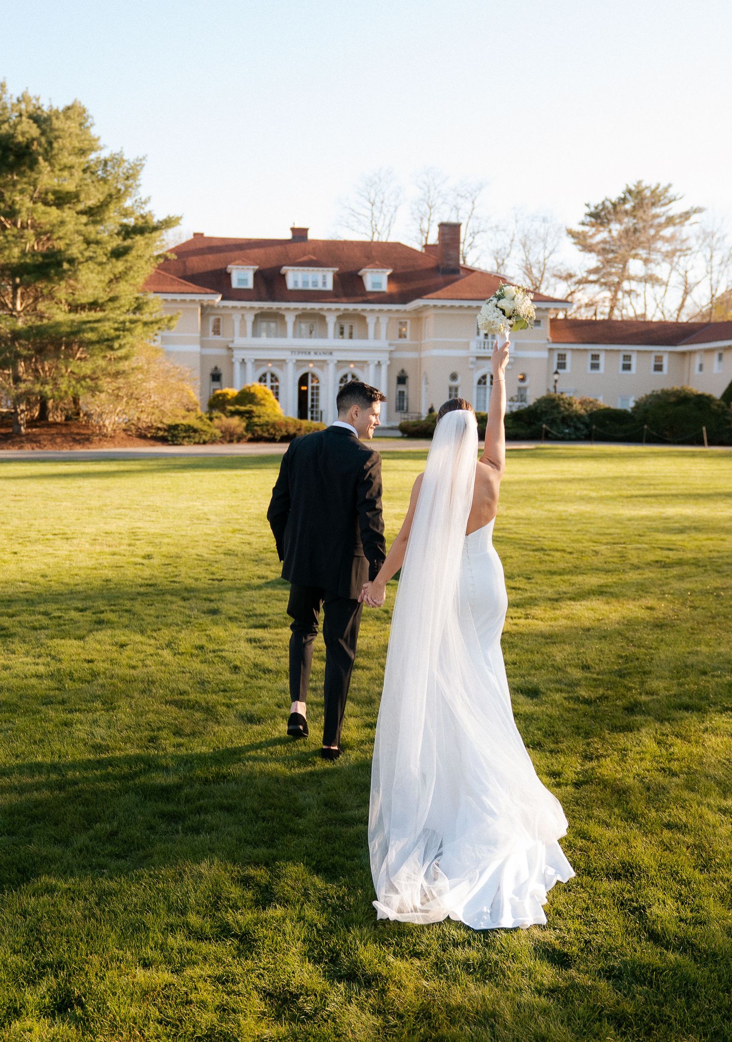 Bride and groom walk hand-in-hand across green lawn toward elegant white mansion wedding venue.