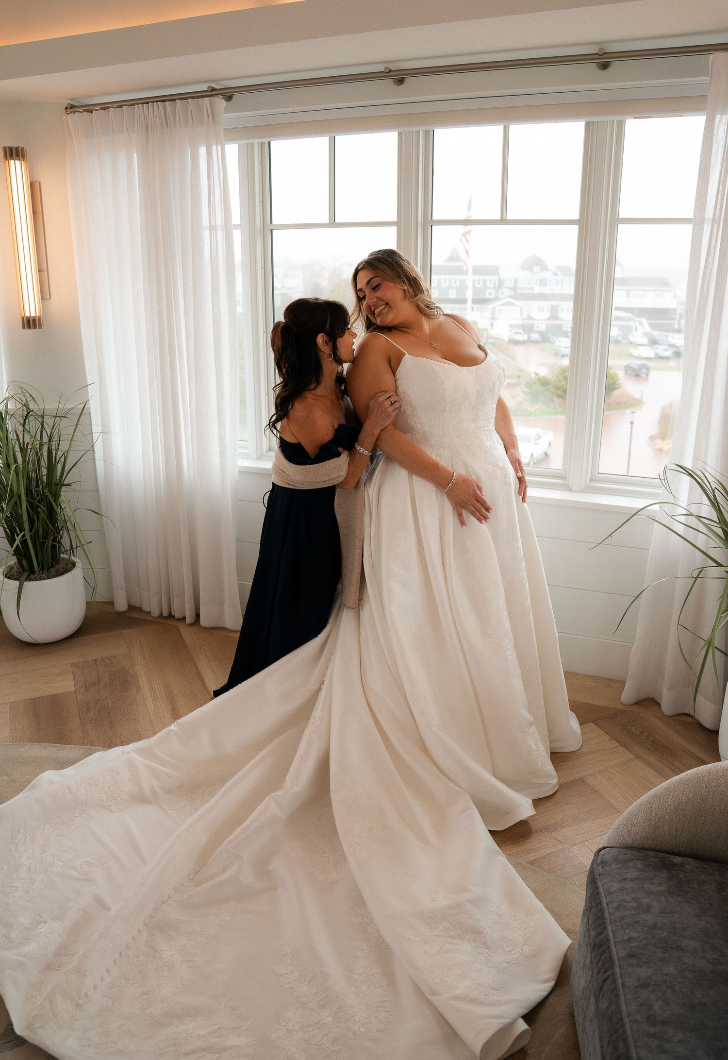 Bride in white ball gown shares tender embrace with bridesmaid in navy dress by sunlit windows.