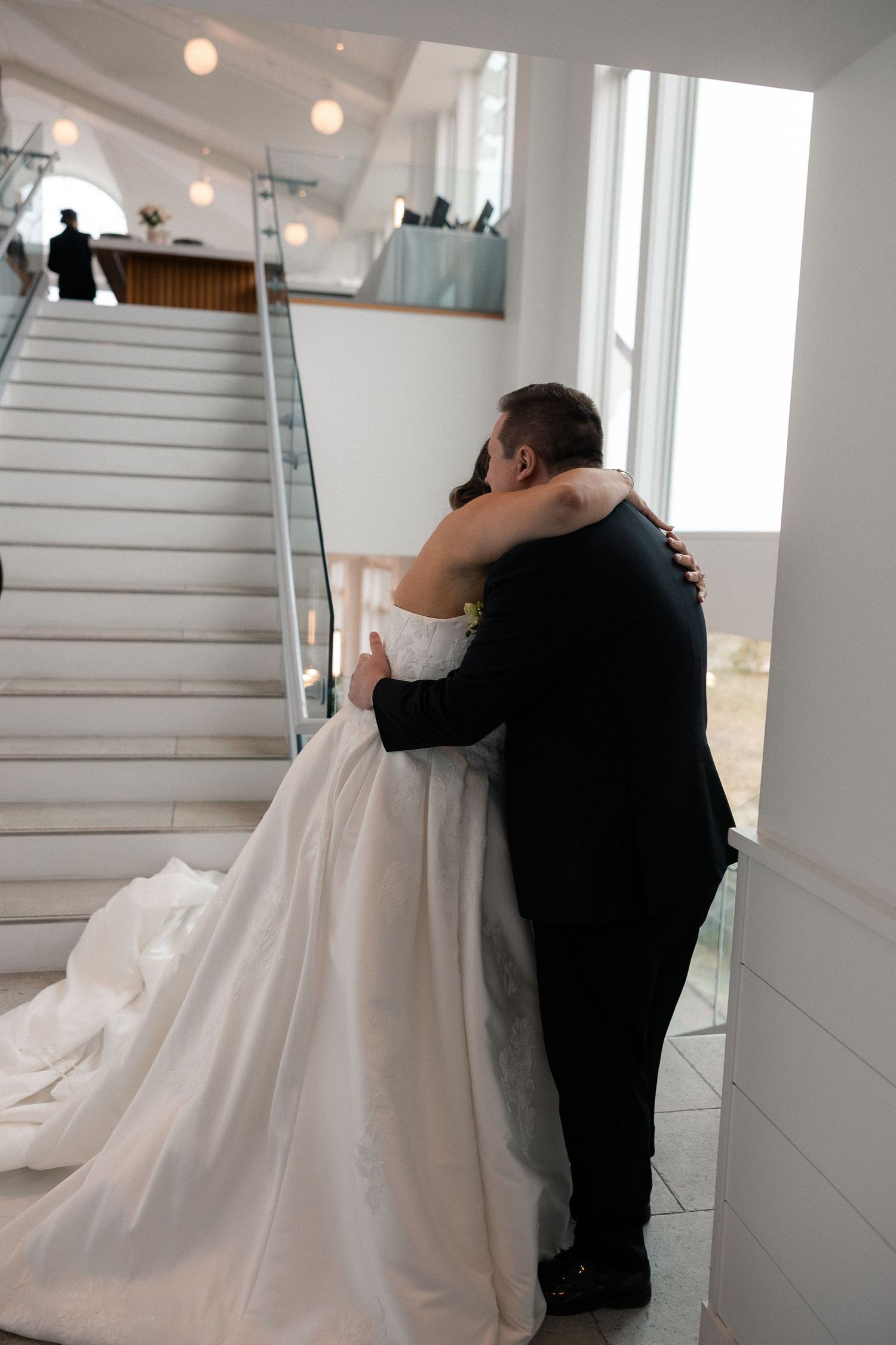 Bride and groom share a tender embrace near elegant staircase, bride in flowing white ball gown at wedding venue.