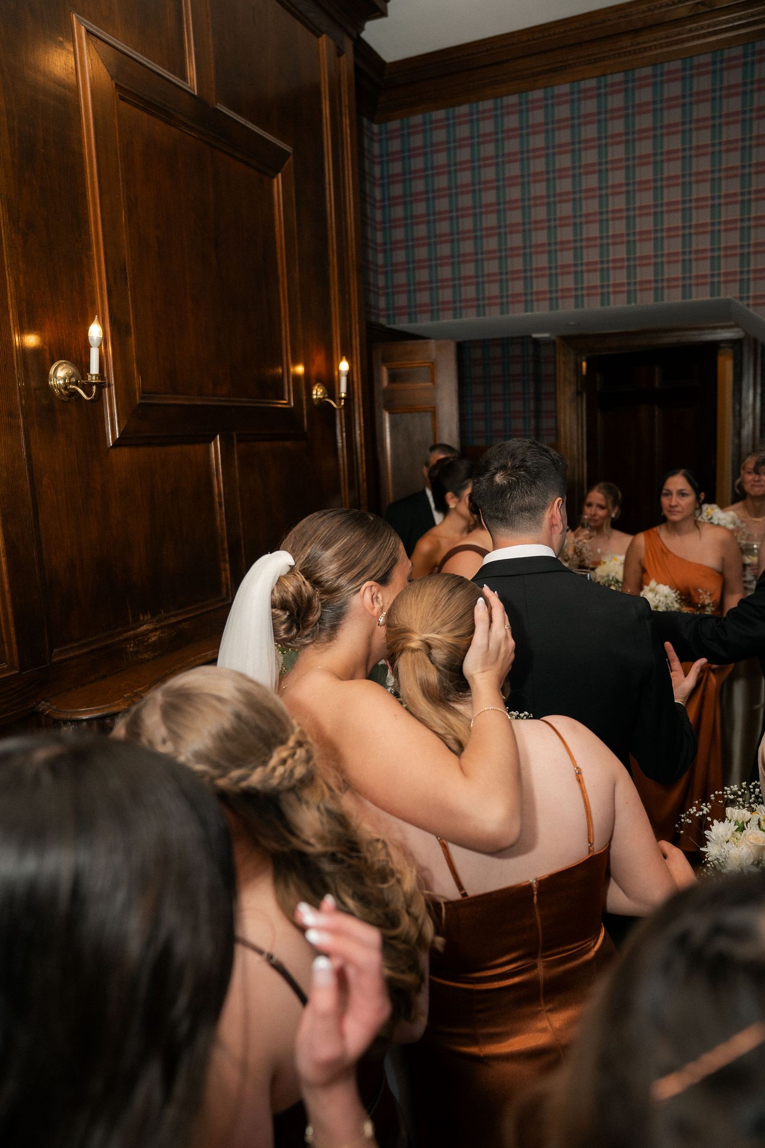 Wedding guests gather in an elegant wood-paneled venue, bridesmaids in rust gowns visible as couple embraces.