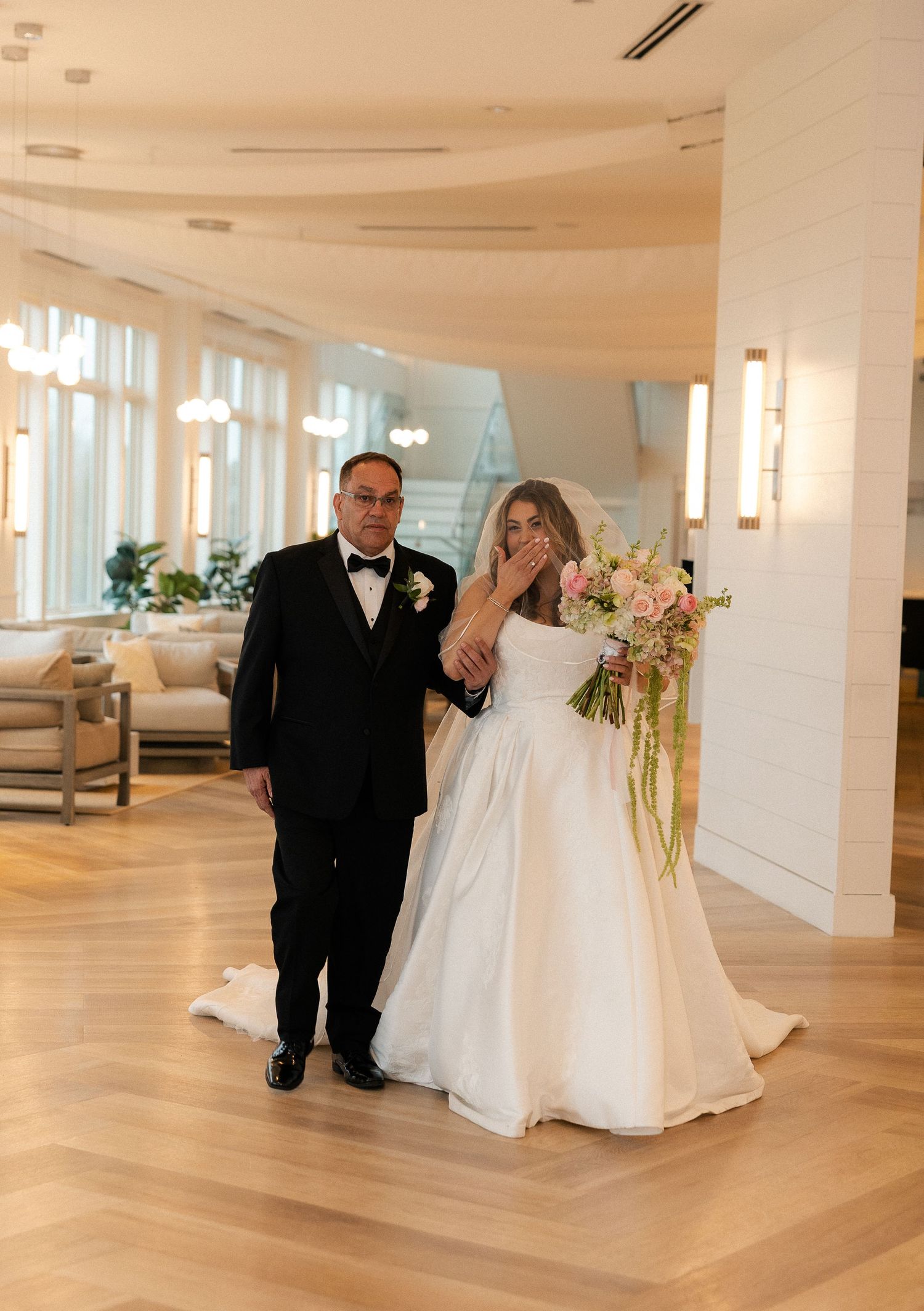 Emotional bride covering her mouth while being escorted by a man in a tuxedo through an elegant venue.