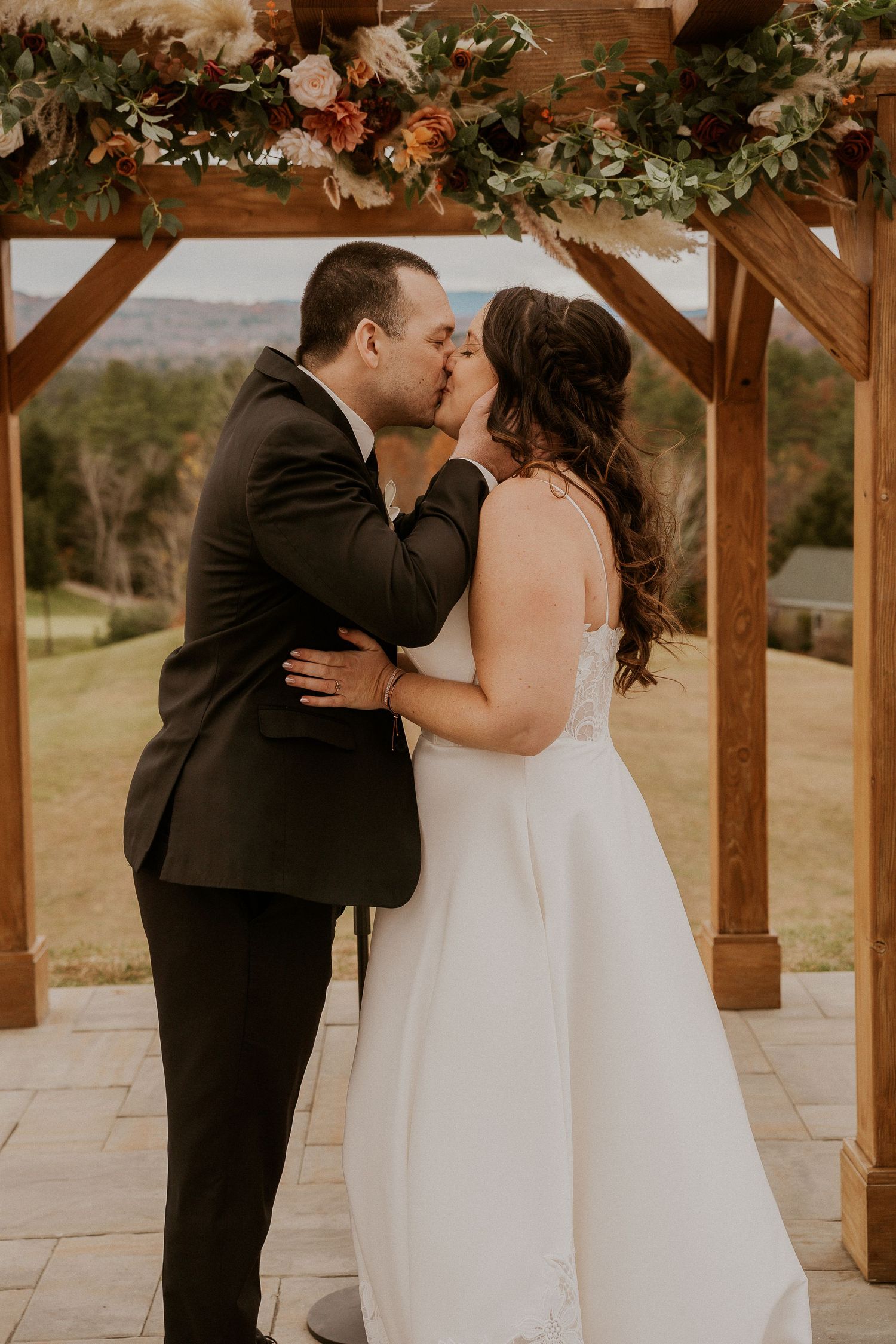 Newlyweds share their first kiss under a wooden arch adorned with greenery and flowers.