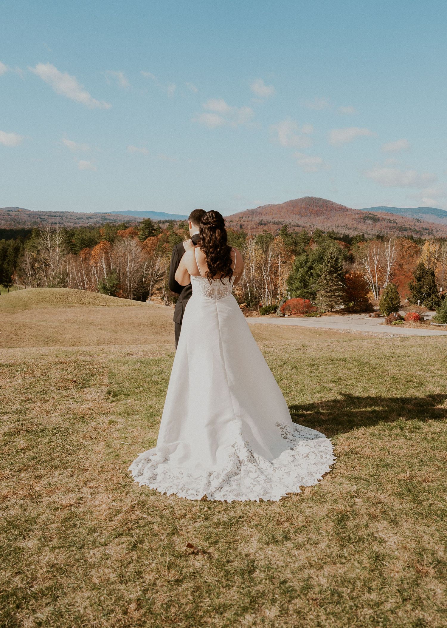 A white wedding dress with lace details flows across a grassy hilltop with mountains and autumn trees in the background.