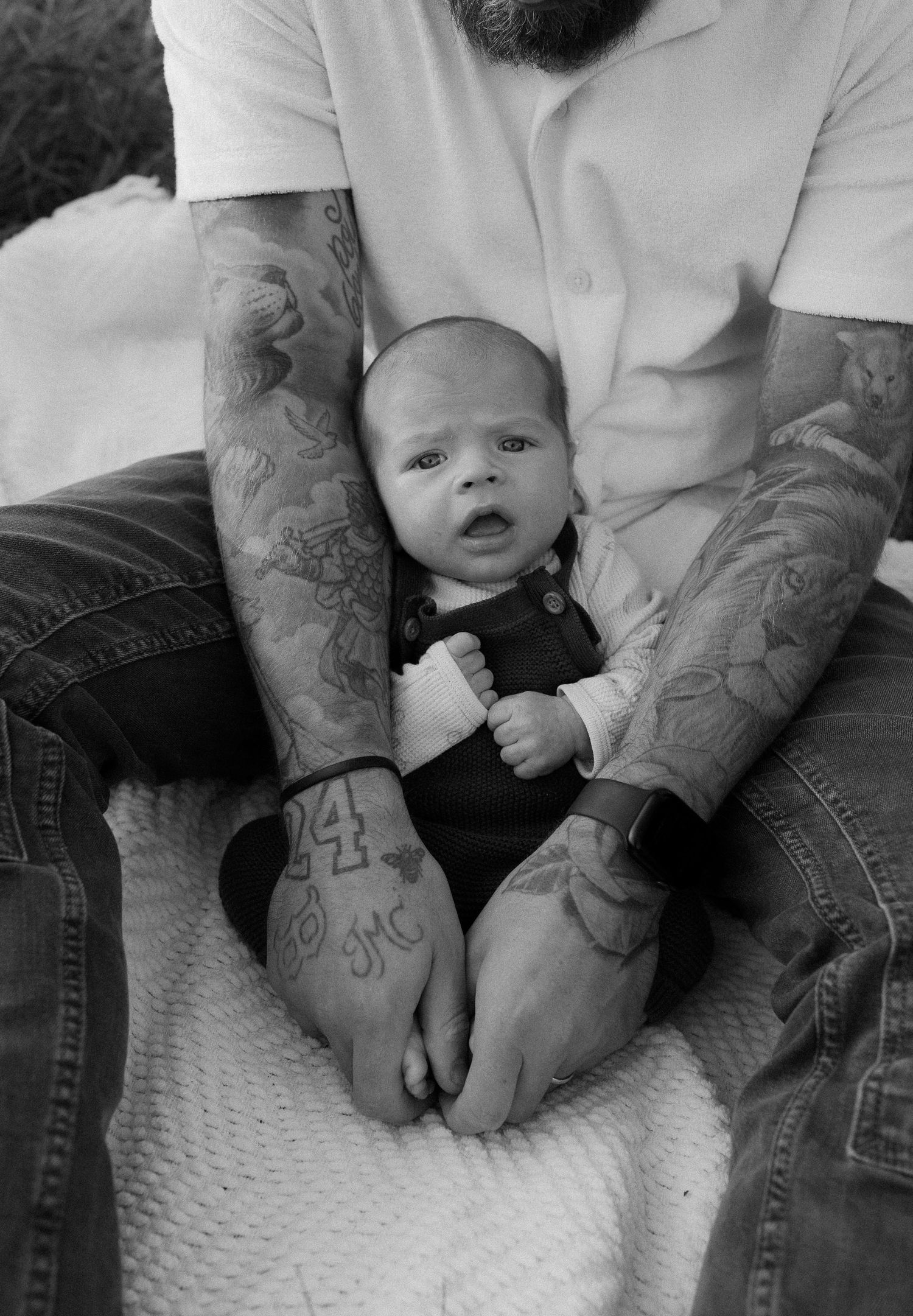 Close-up view of tattooed arms holding a baby on a white blanket.