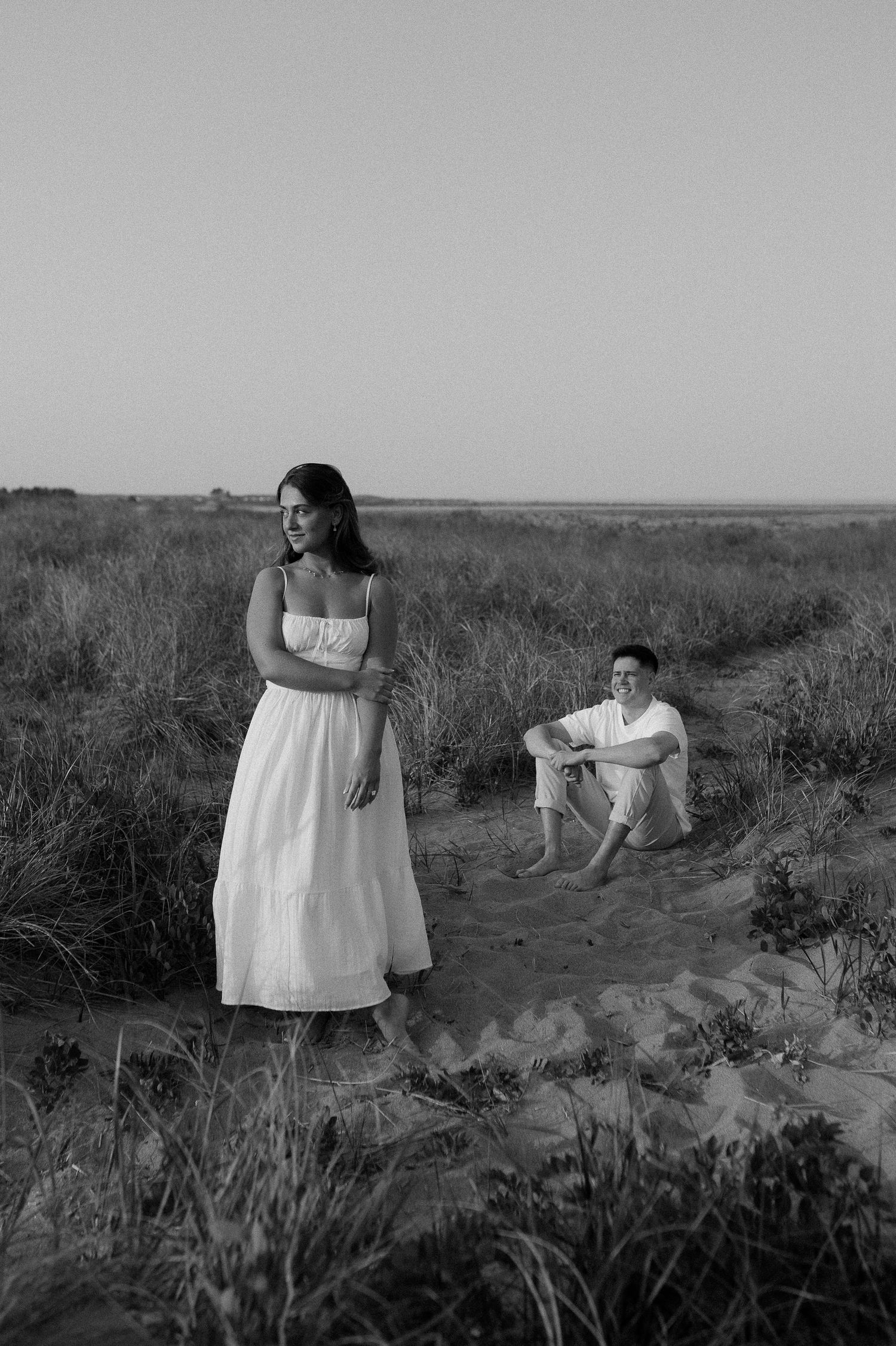 Couple in black and white photo posing on sandy dunes with natural grassland backdrop.