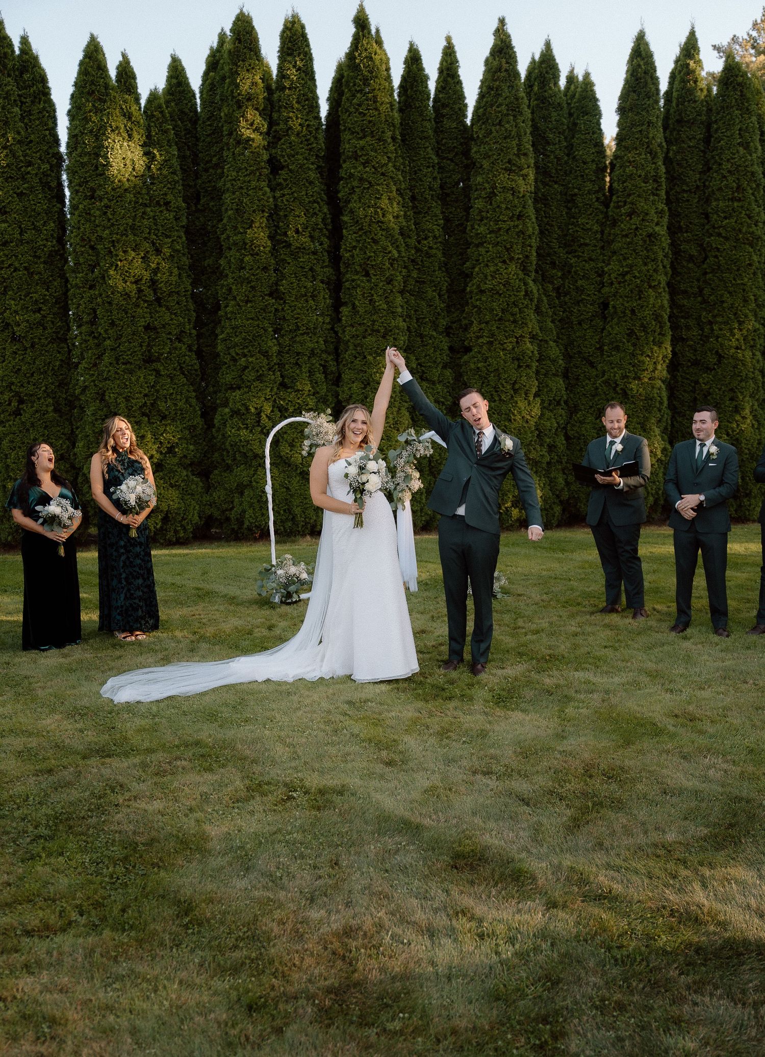 Wedding guests stand in formal attire on a grassy lawn lined with tall evergreen trees.