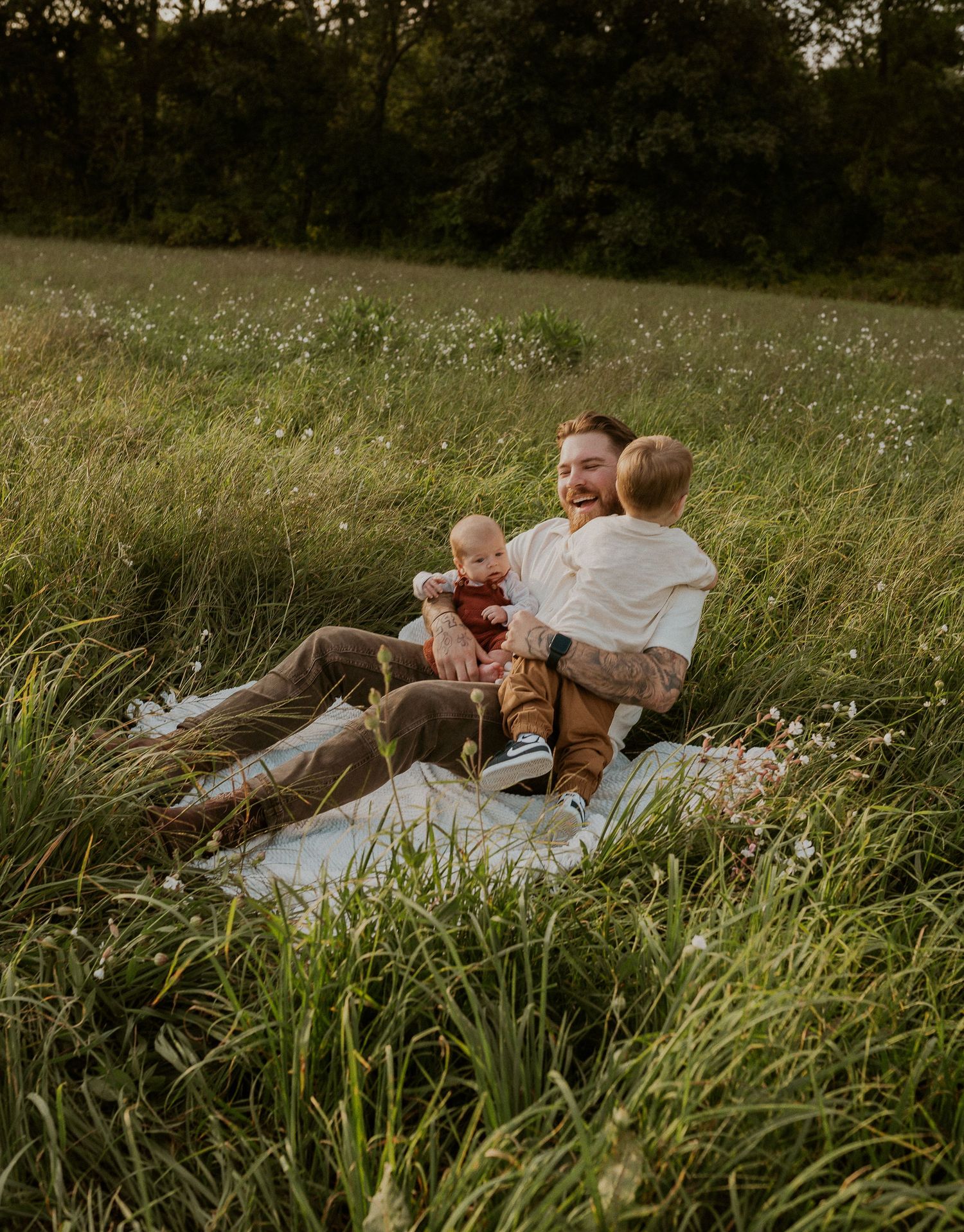 Family lying together on white blanket surrounded by tall grass at sunset.