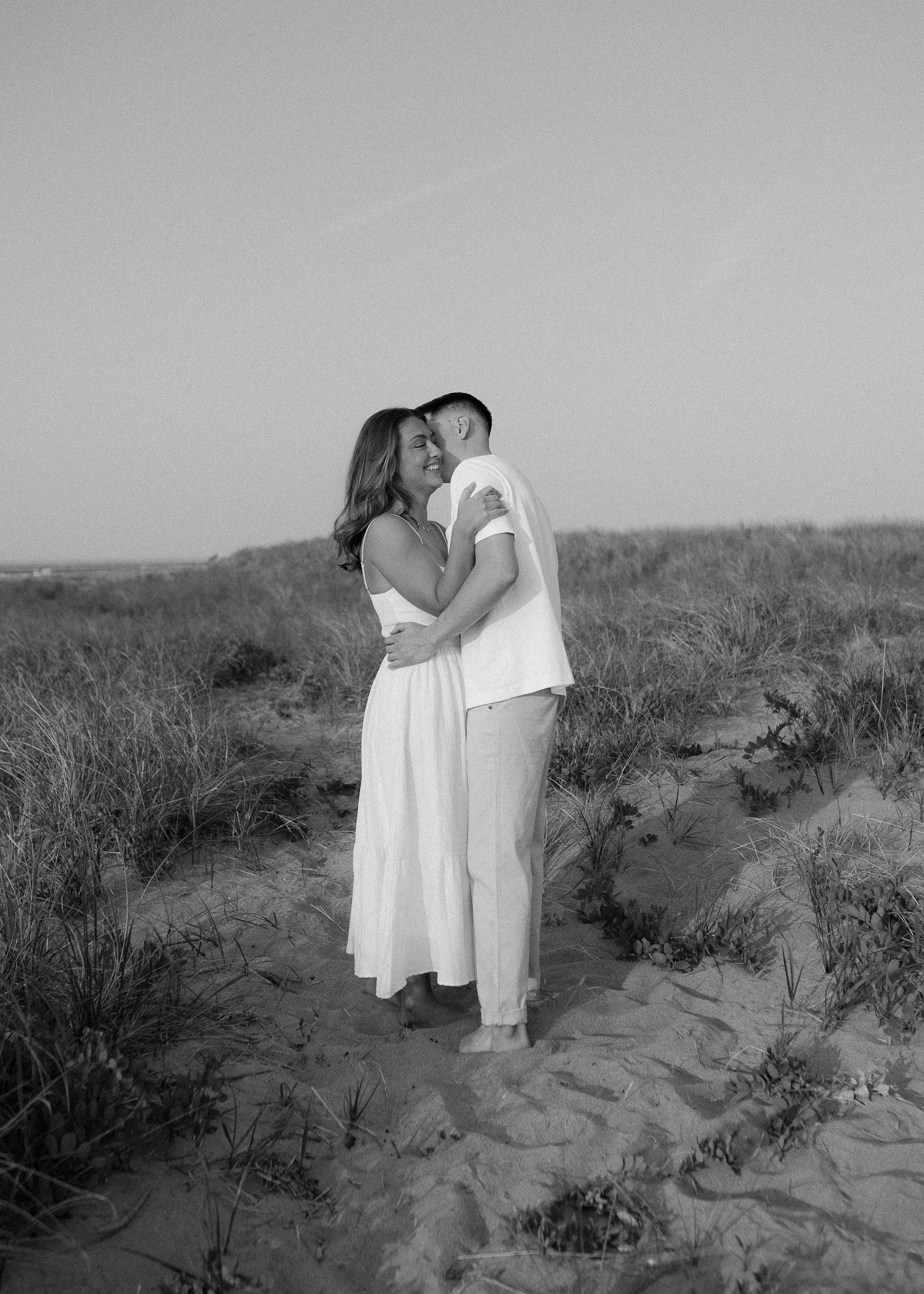 Black and white landscape photos of a couple embracing on sandy dunes with wild grass.