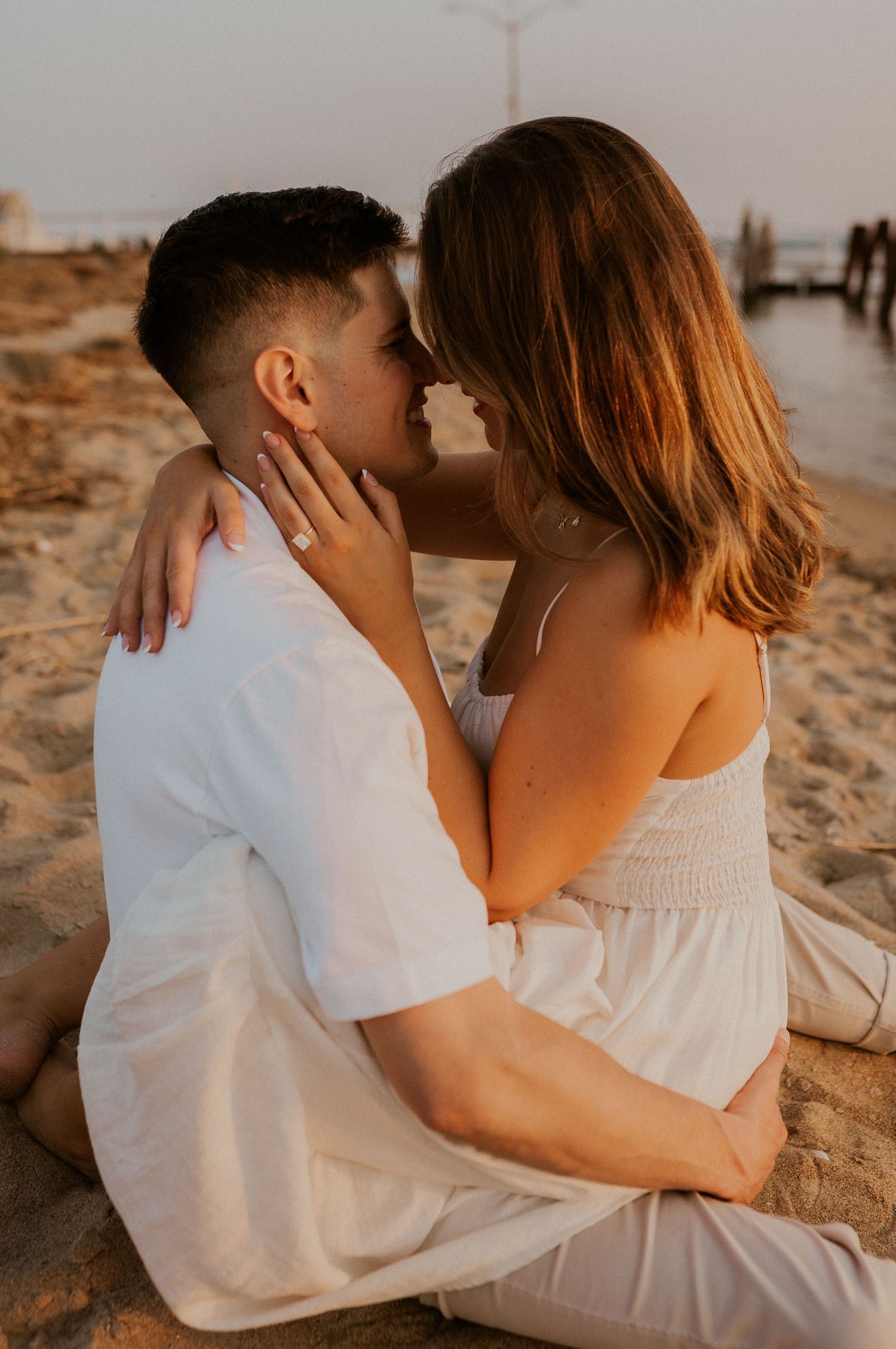 Couple in white clothing sitting close together on beach sand during golden hour.