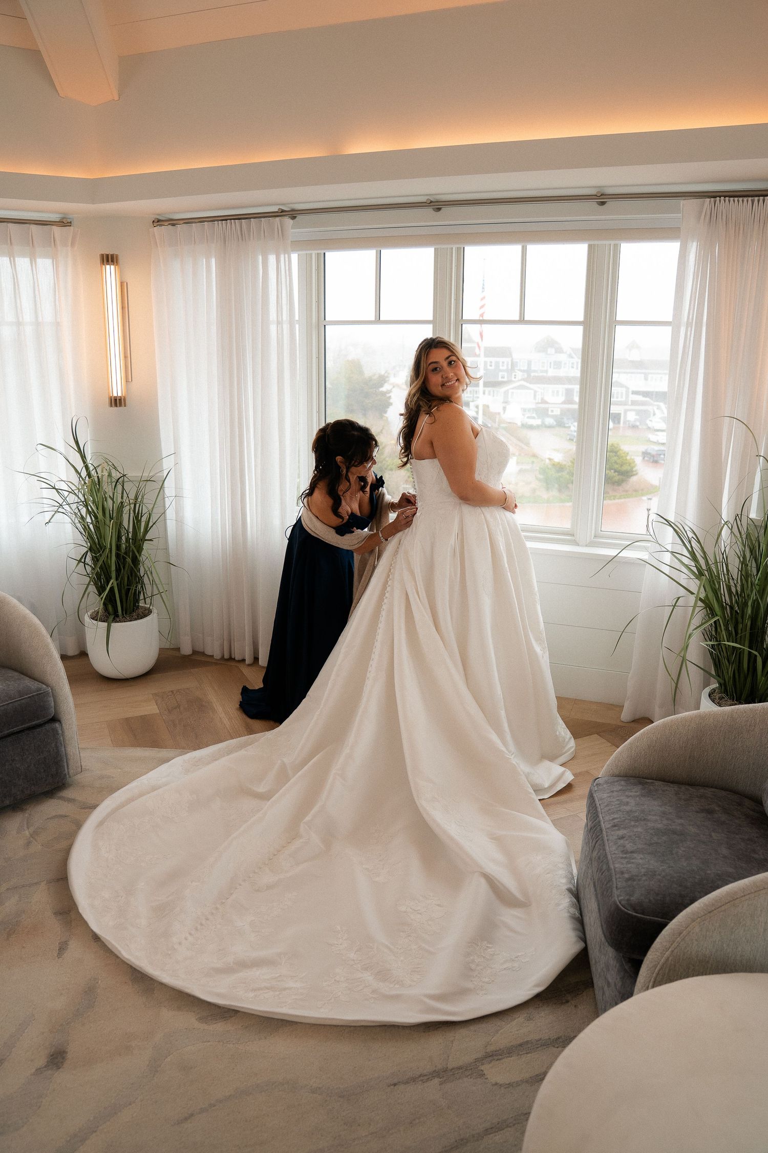 Bride in elegant white ball gown with long train being helped by bridesmaid near bright windows in a modern bridal suite.