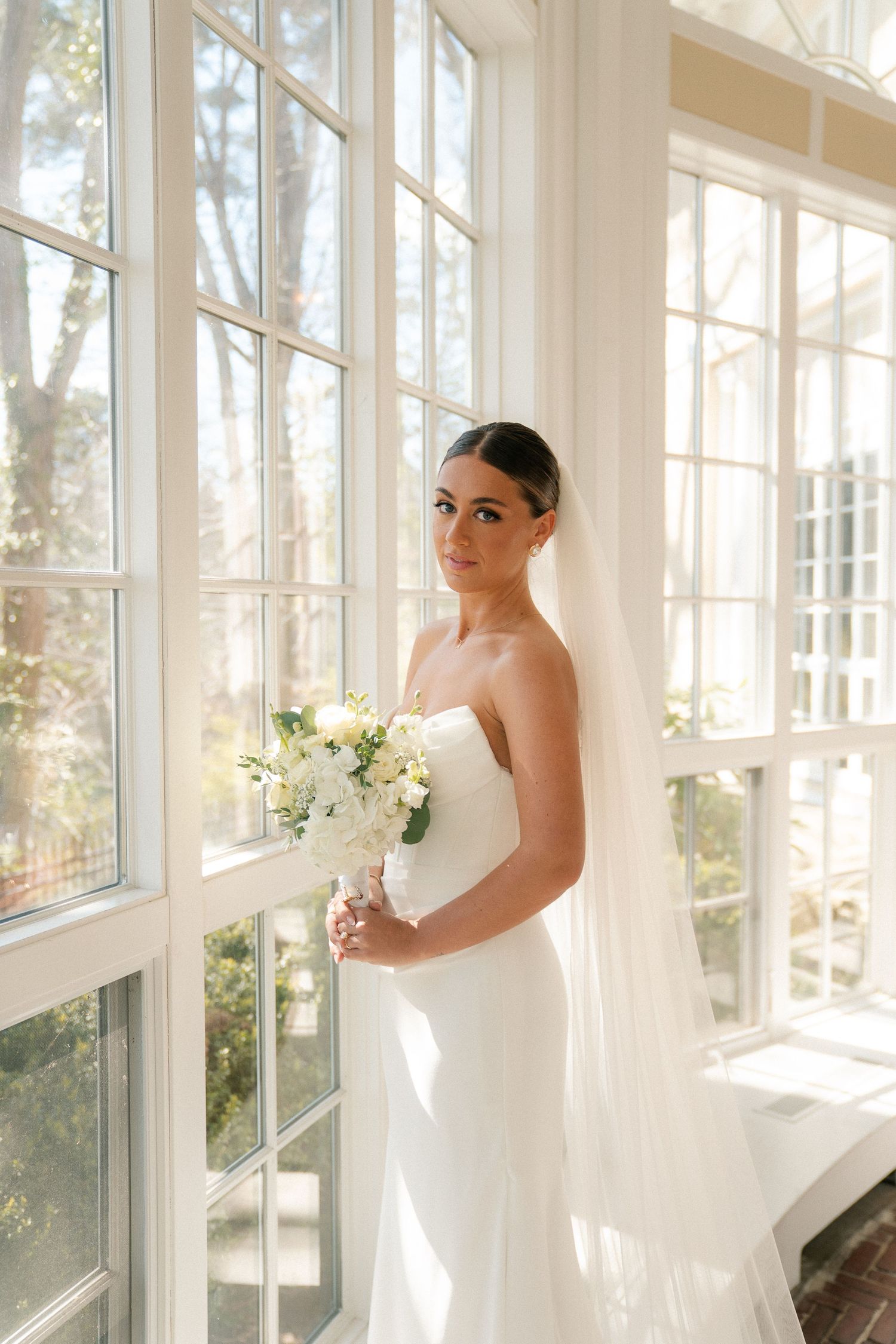 Elegant bride in strapless white gown holding floral bouquet, standing by large sunlit windows with veil flowing.