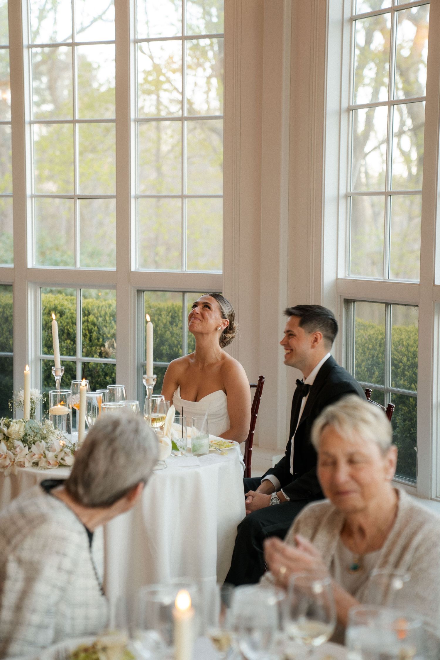 Bride and groom laugh at wedding reception table, seated before large windows with candles and floral centerpieces.