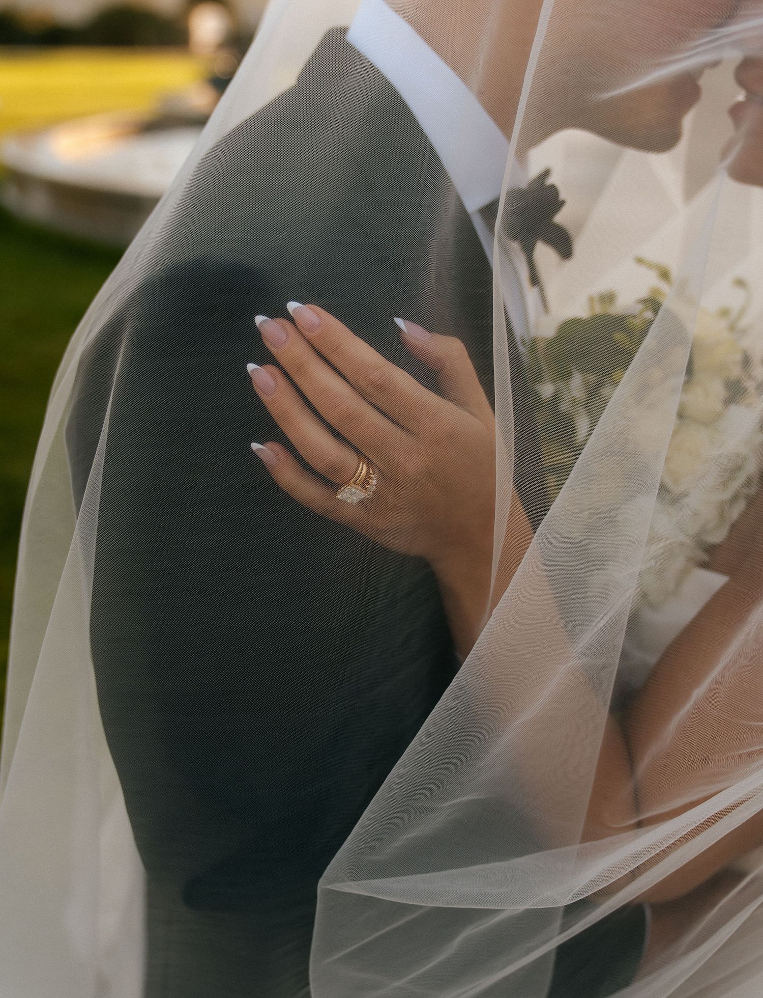 Bride and groom embrace under a sheer wedding veil, showcasing her manicured nails and gold wedding band.