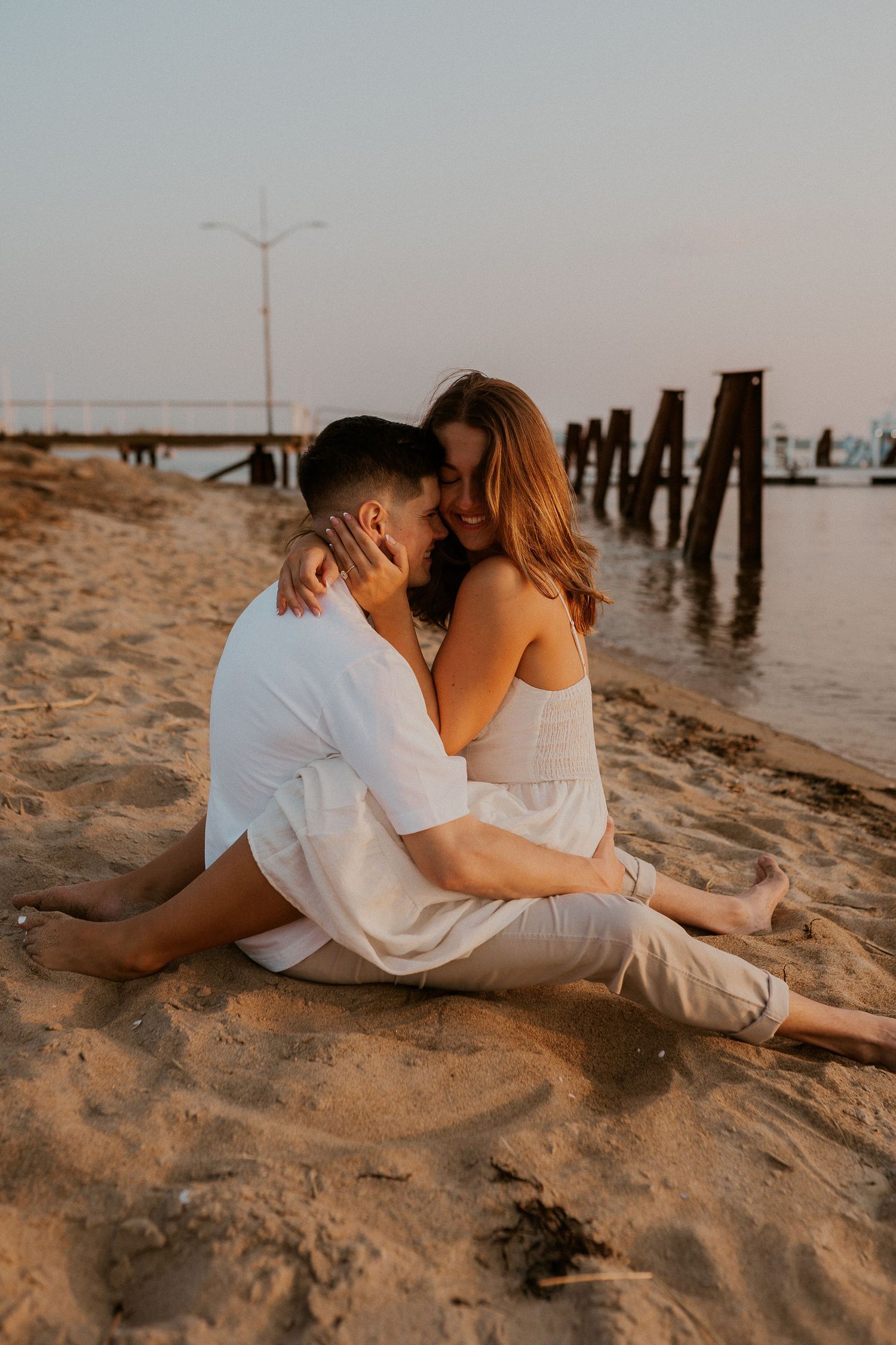 Intimate beach portrait of couple sitting on sand near wooden pier posts at sunset.