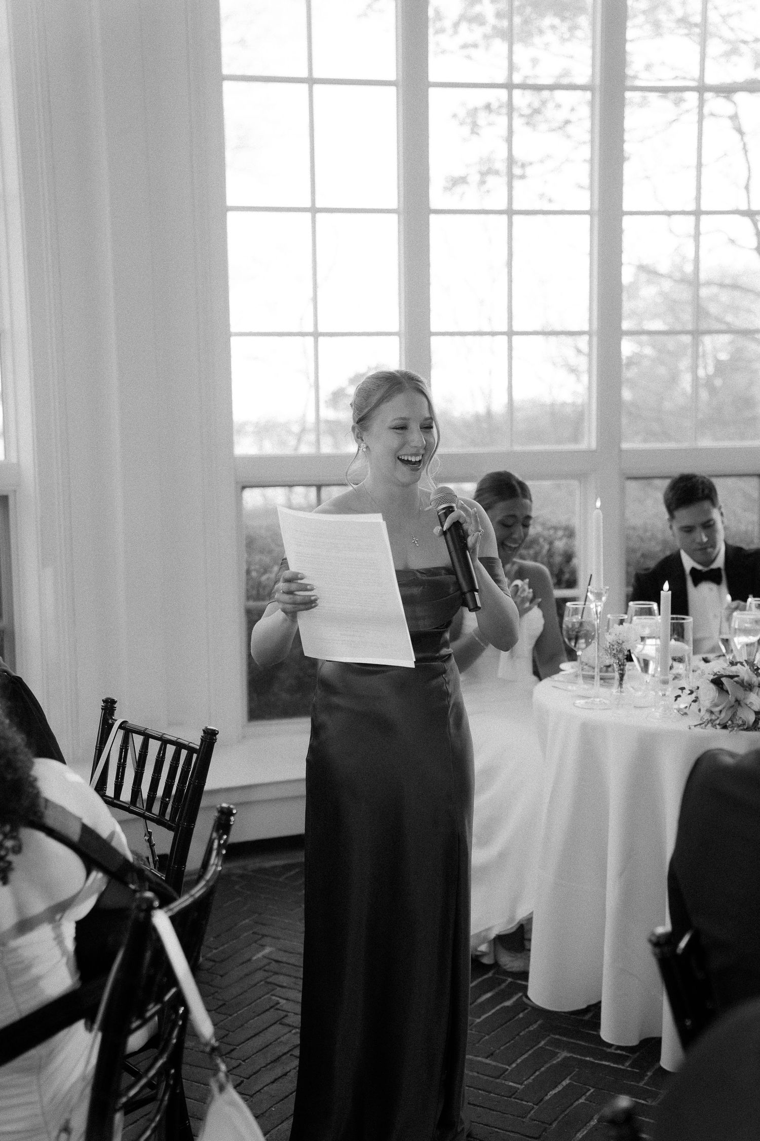 Bridesmaid in elegant gown delivers heartfelt speech at wedding reception, holding paper and microphone.