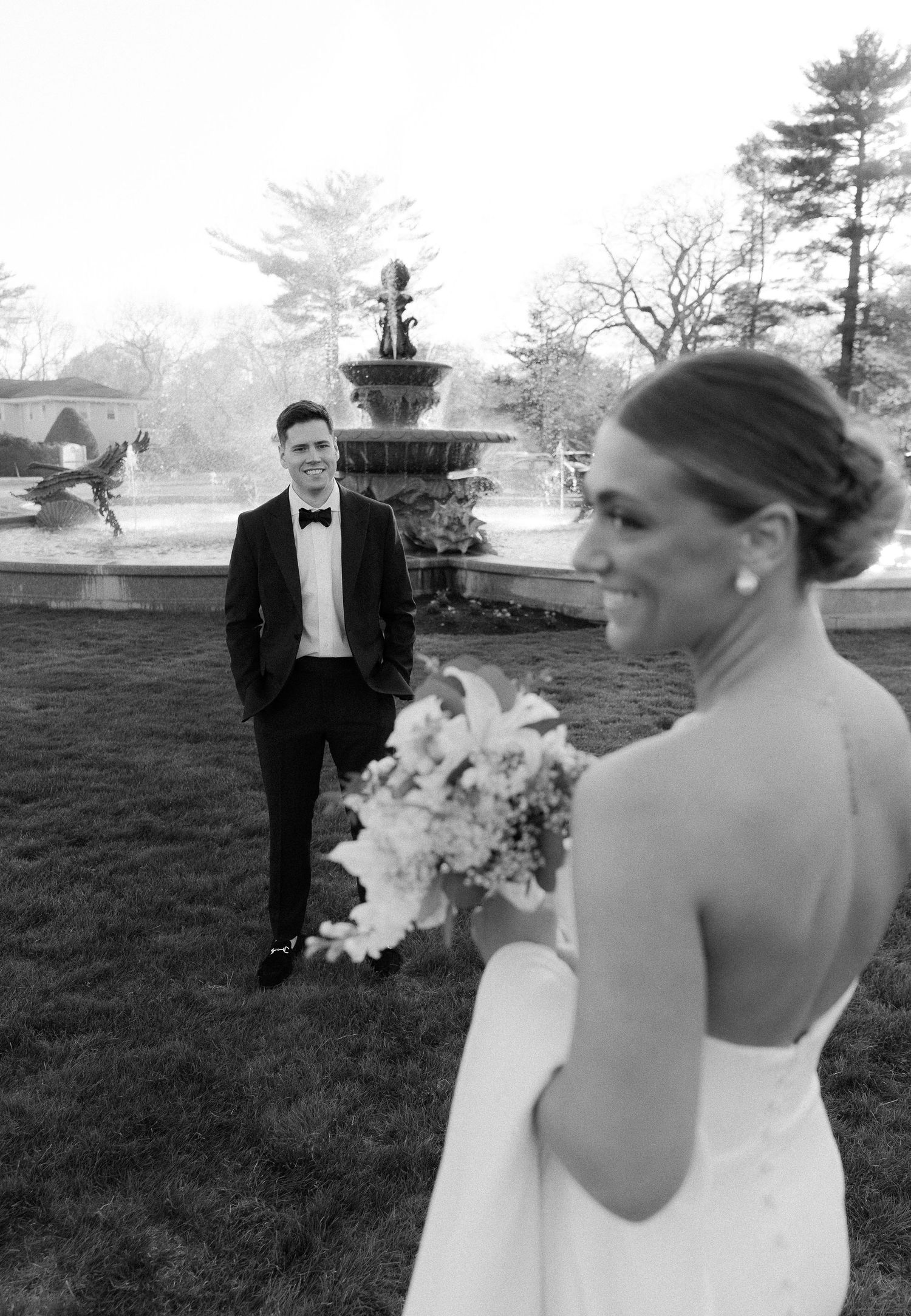 Groom in tuxedo smiles at bride holding bouquet during first look near elegant stone fountain.