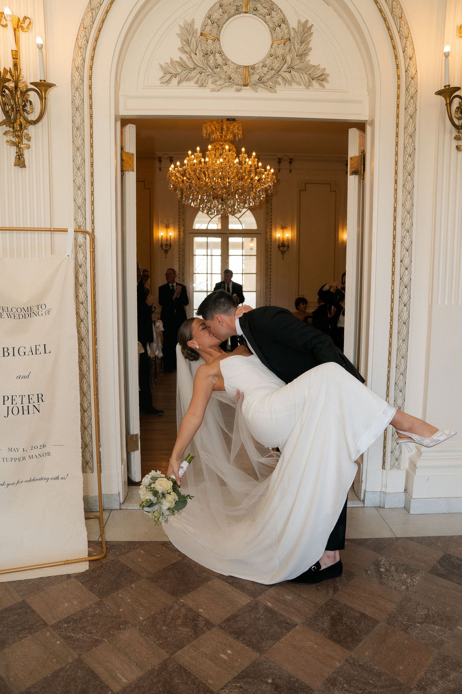 Groom dips bride for a passionate kiss in an elegant arched doorway with chandelier glowing behind them.