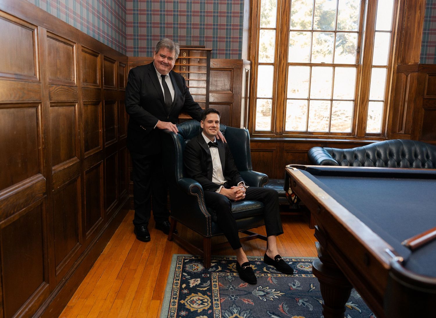 Two men in formal tuxedos posing in a wood-paneled billiard room, one seated in leather chair.