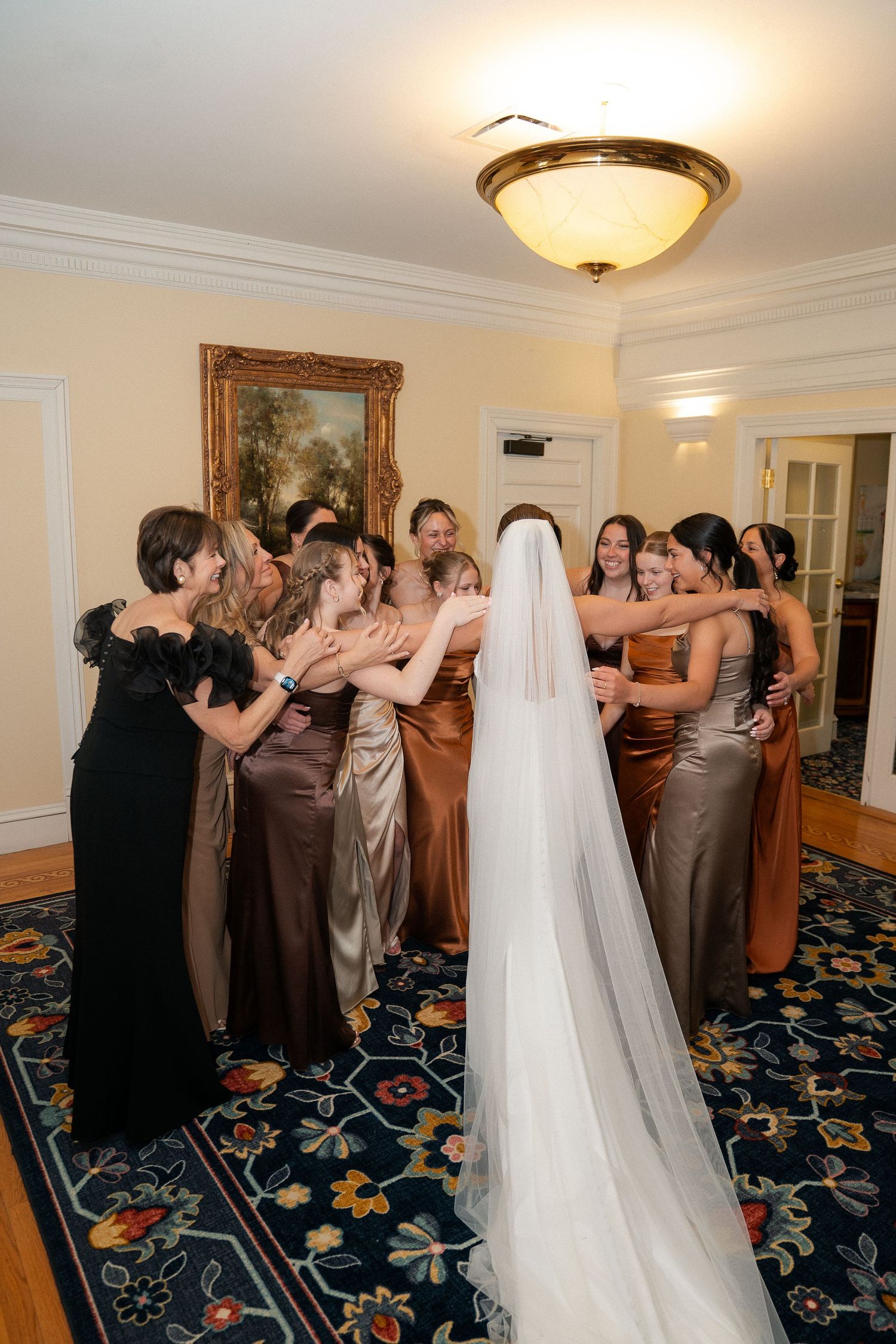 Bride in white veil surrounded by bridesmaids in brown satin gowns in elegant room during wedding preparations.