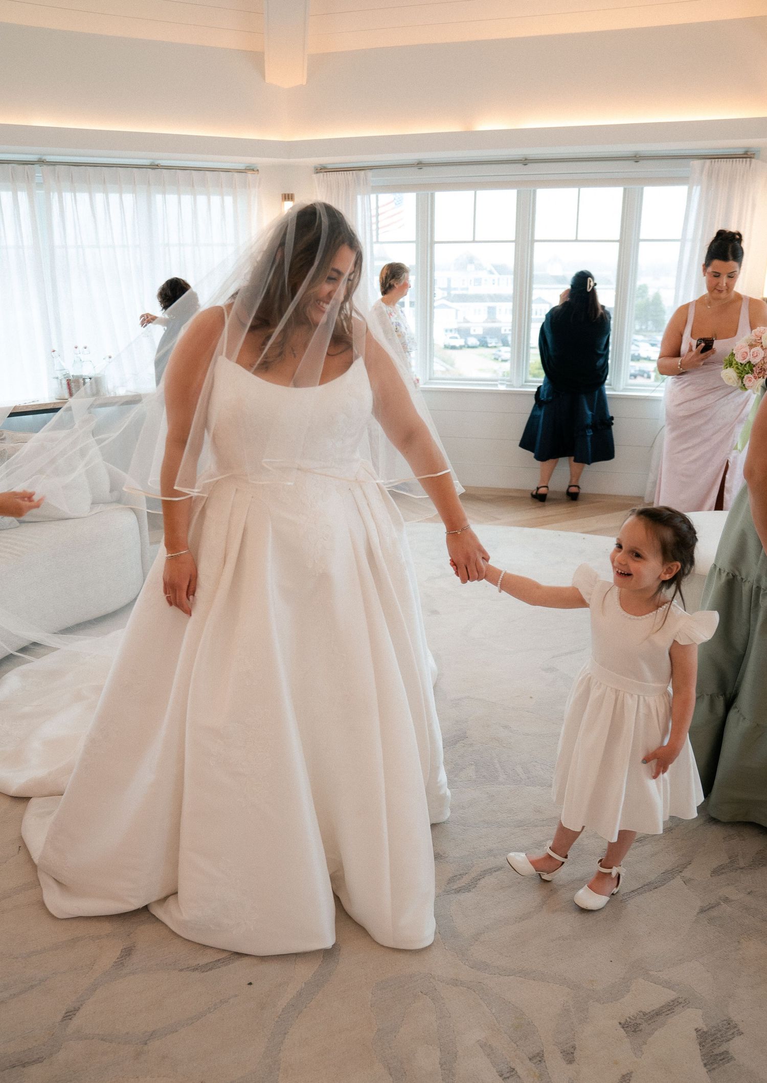 Bride in white ball gown and veil holds hands with young flower girl in white dress during wedding preparations.