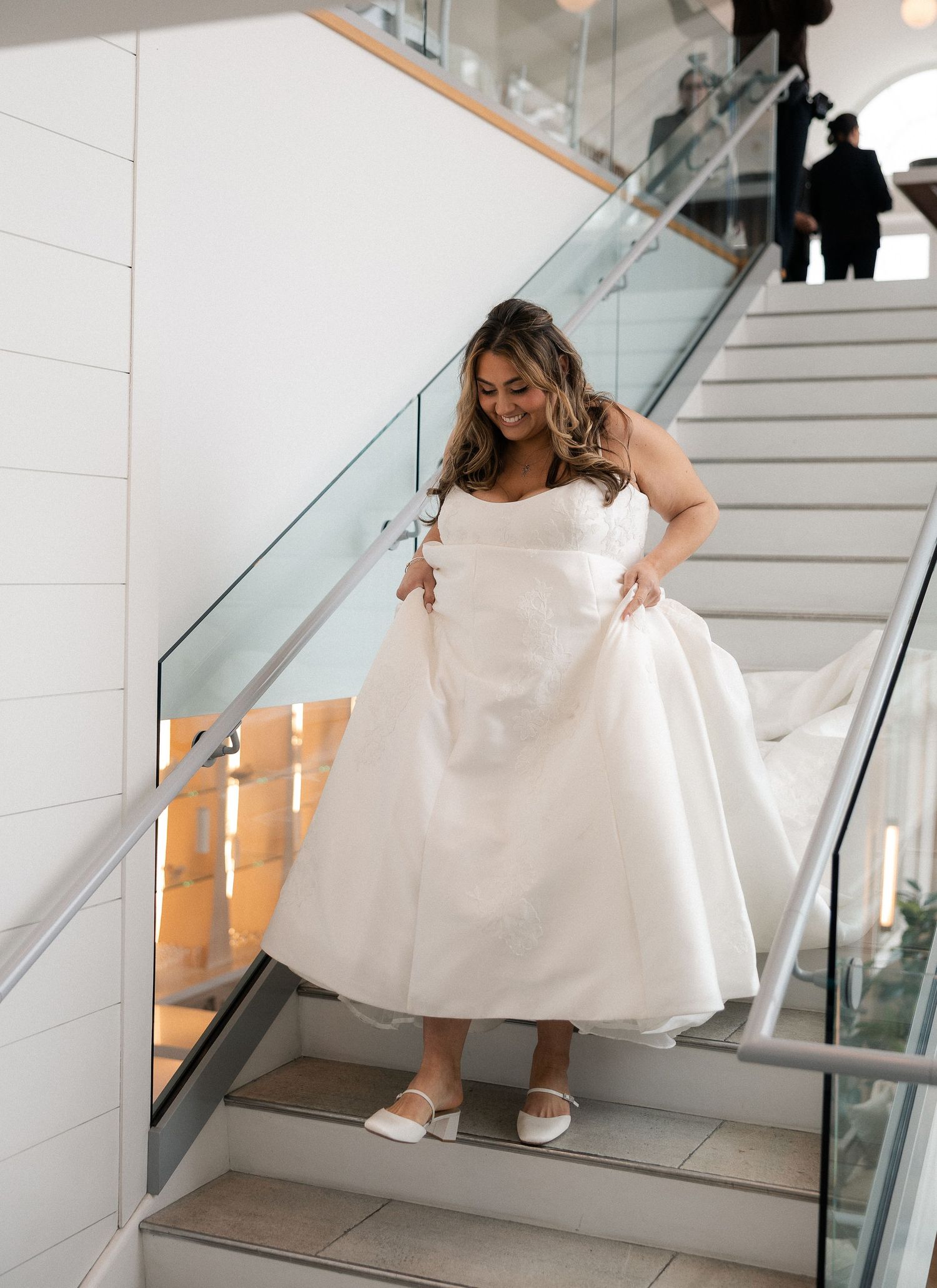 Smiling bride in white tea-length wedding dress walking down elegant staircase.
