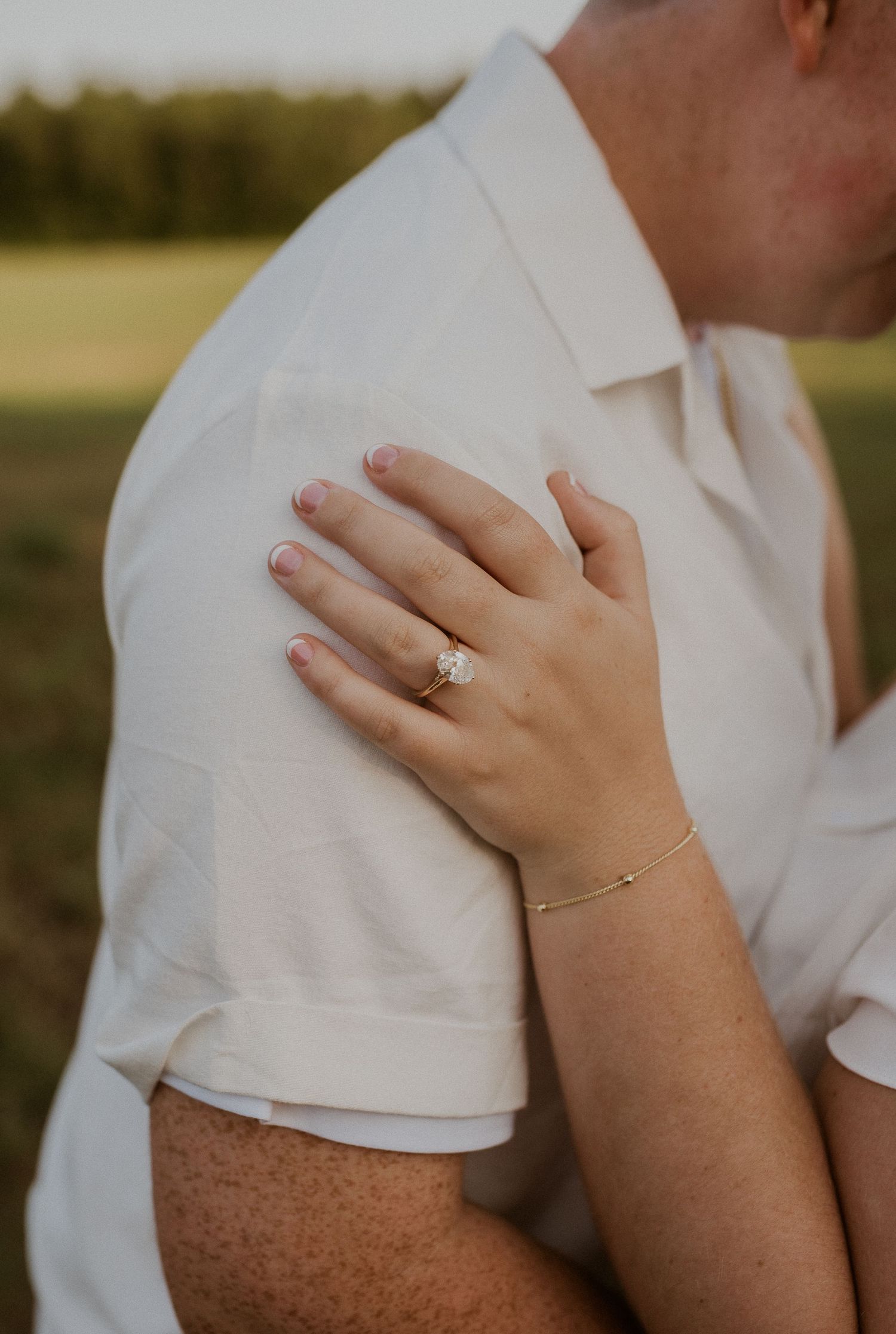 A close-up view of hands wearing an engagement ring and delicate bracelet against a white shirt.