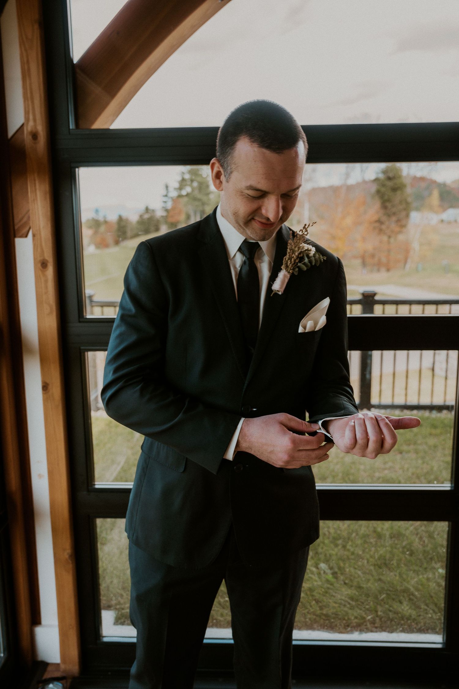Person in a black suit checking their watch while standing near a window.