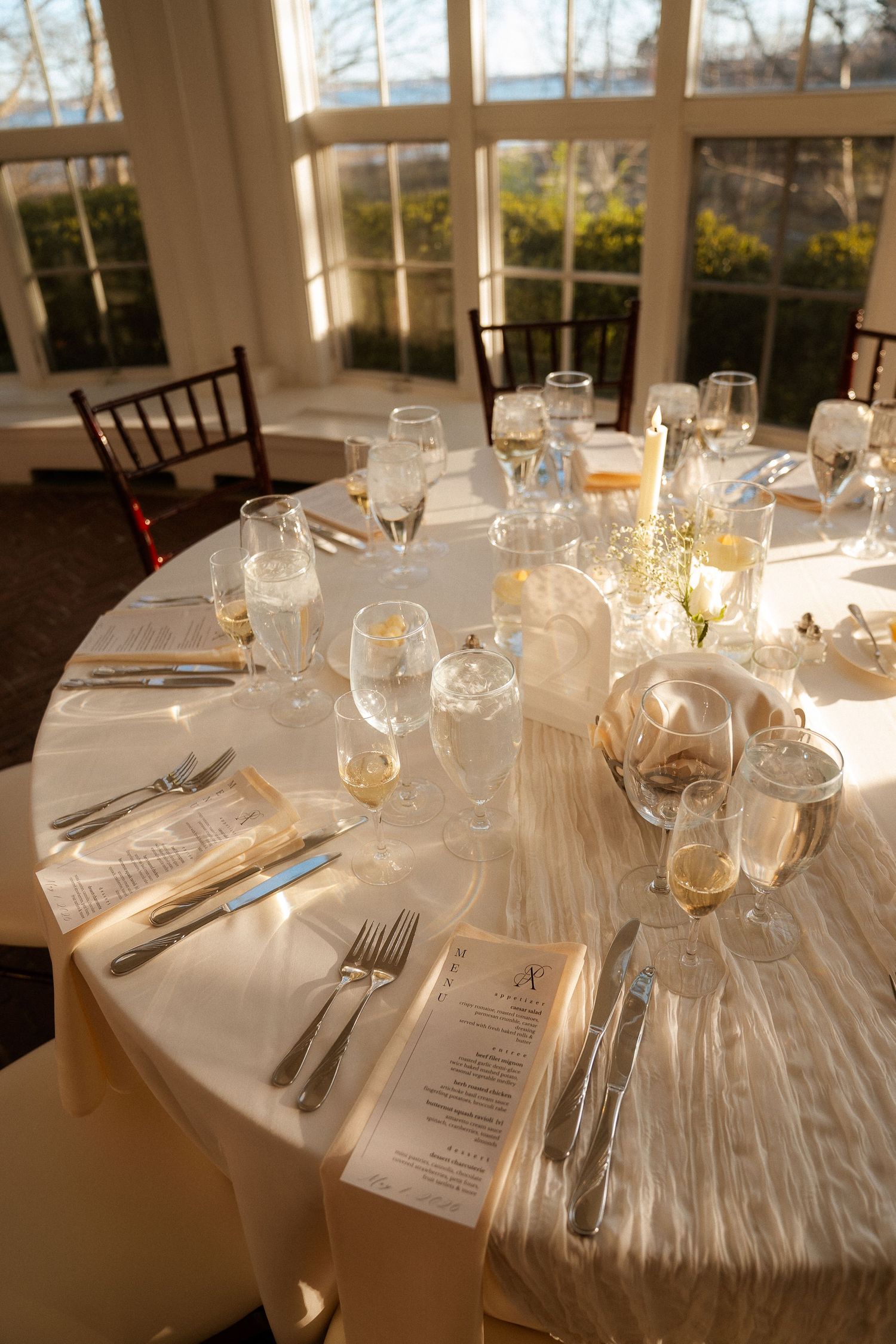 Elegant round wedding reception table with white linens, crystal glassware, and menus in a sunlit venue.