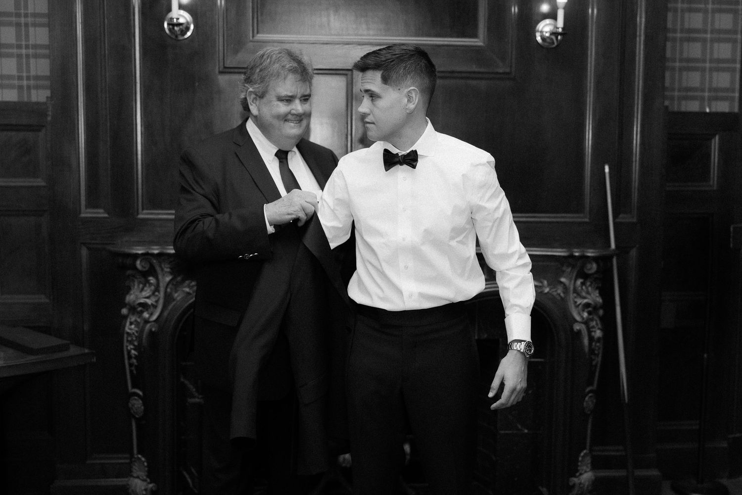 Two men in formal attire share a moment in a classic interior, one in a suit and the other in a tuxedo shirt with bow tie.