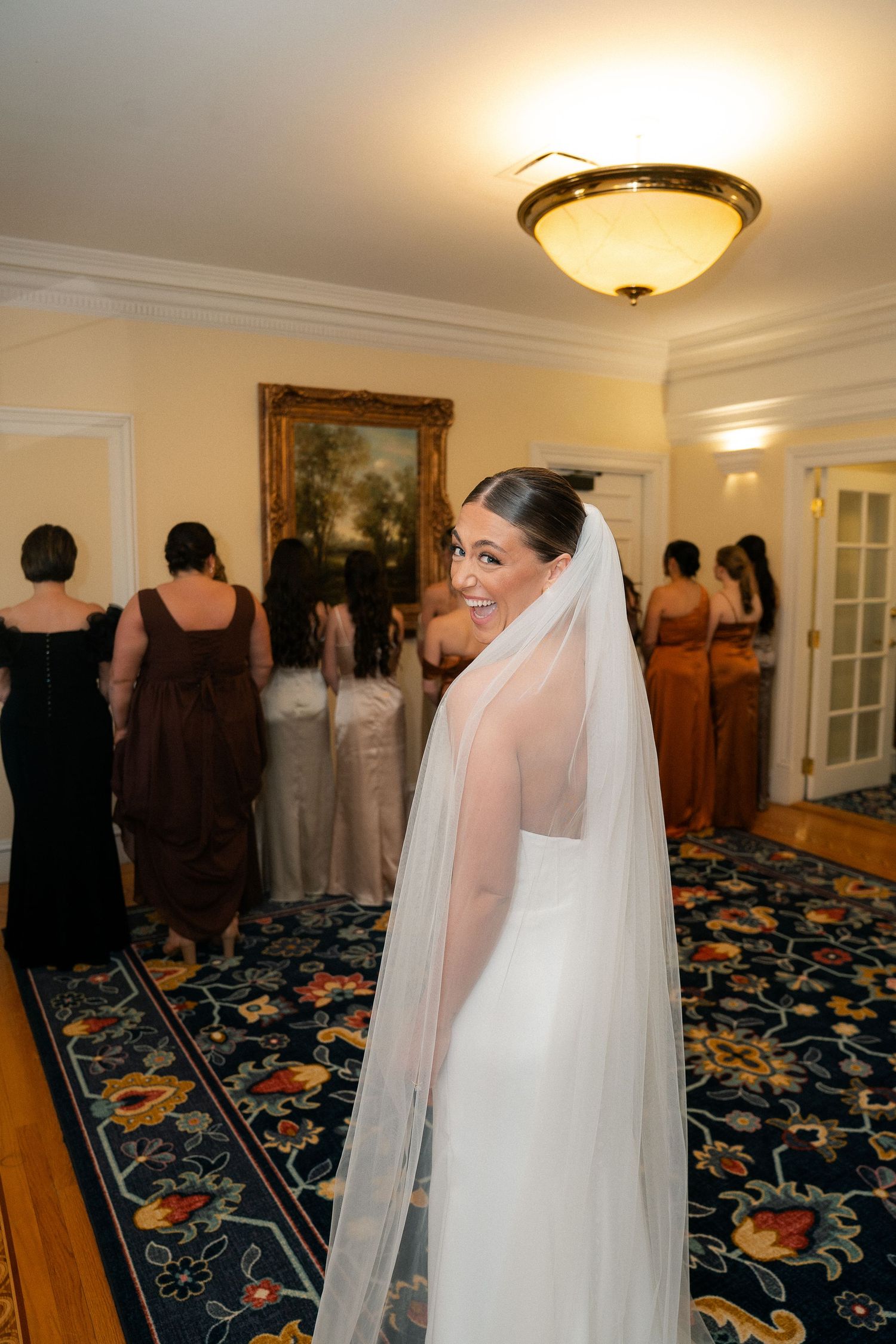 Smiling bride in white gown and veil looks back at camera while bridesmaids stand in background in elegant room.