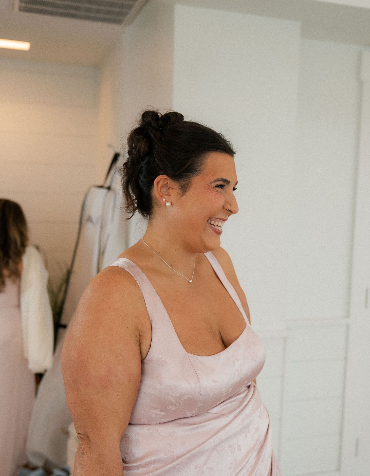 A smiling woman with dark updo hair wearing a blush pink satin dress and delicate jewelry in a bright white room.