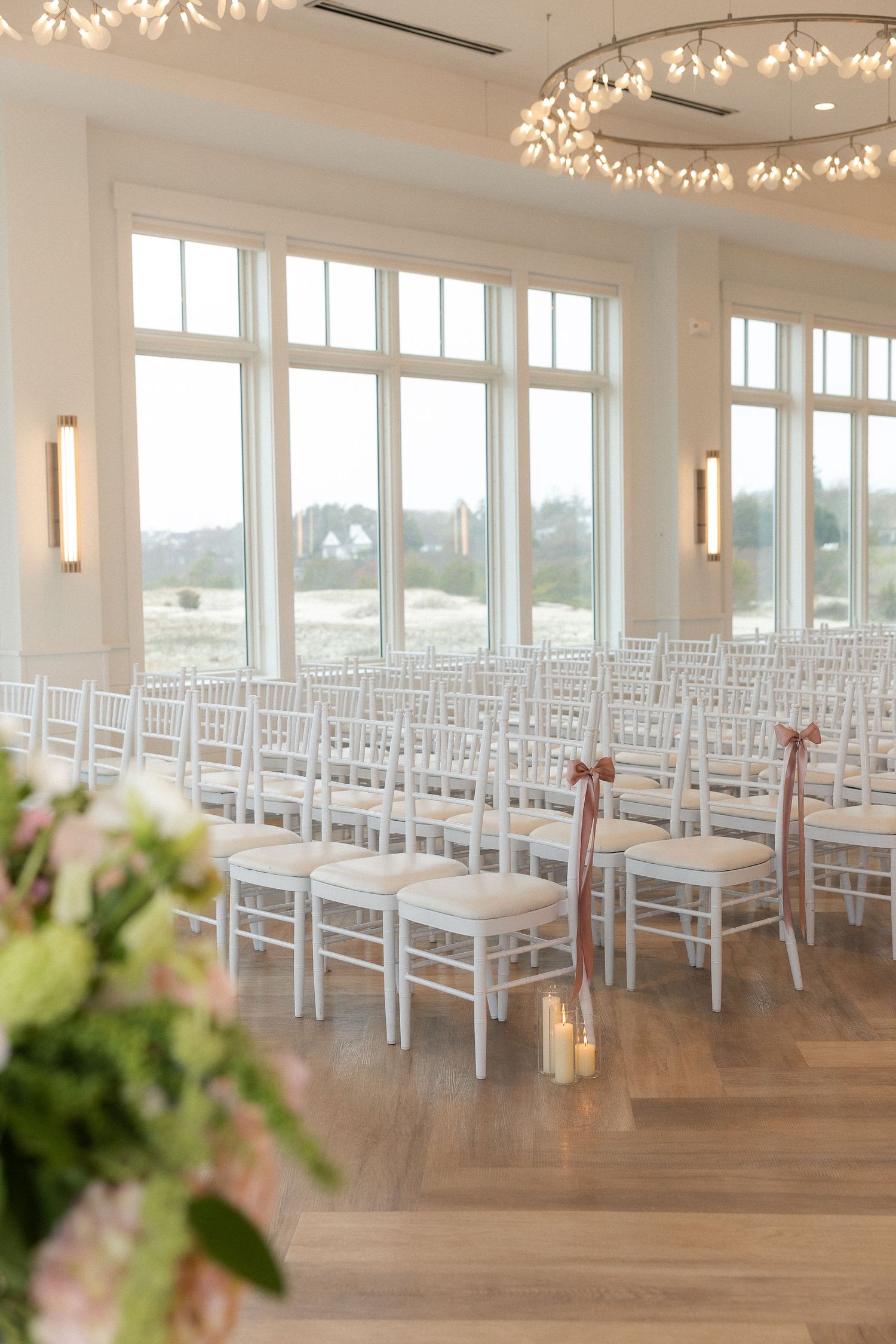 Elegant wedding ceremony venue with white chiavari chairs, blush ribbon accents, large windows, and a circular chandelier.