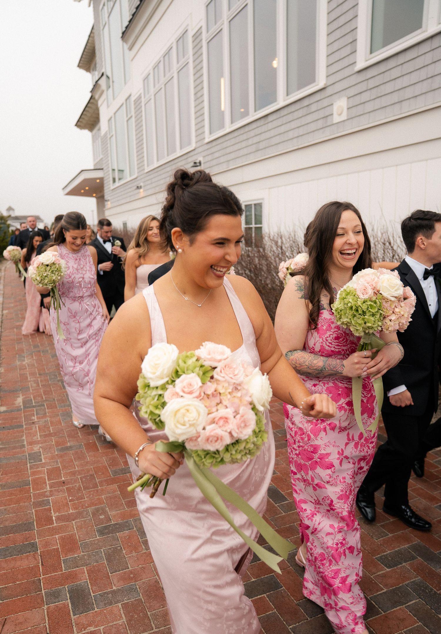 Bridesmaids in pink floral dresses laugh while walking outdoors holding bouquets of roses and green hydrangeas.