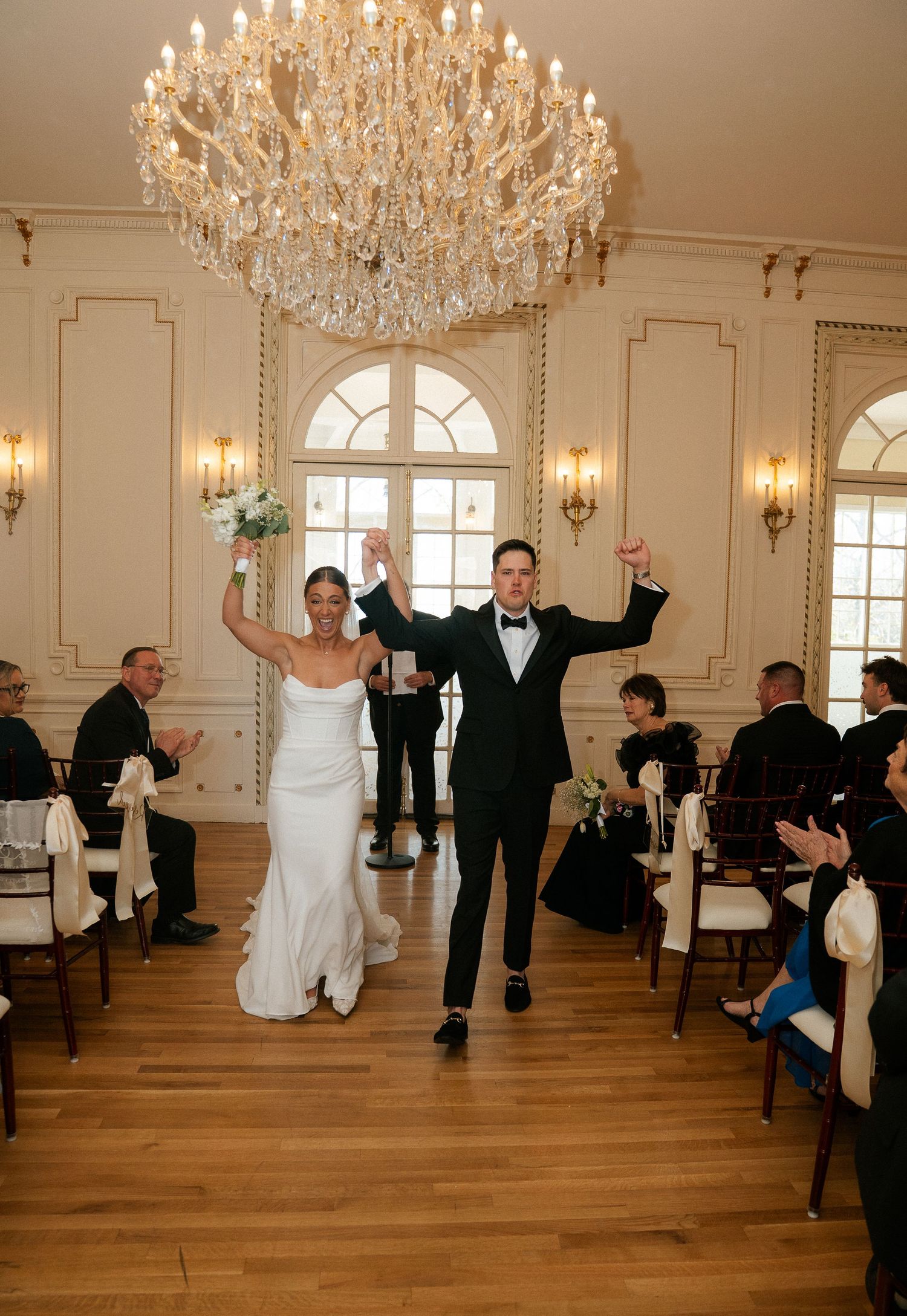 Joyful newlyweds raise hands triumphantly while walking down the aisle beneath a grand crystal chandelier.