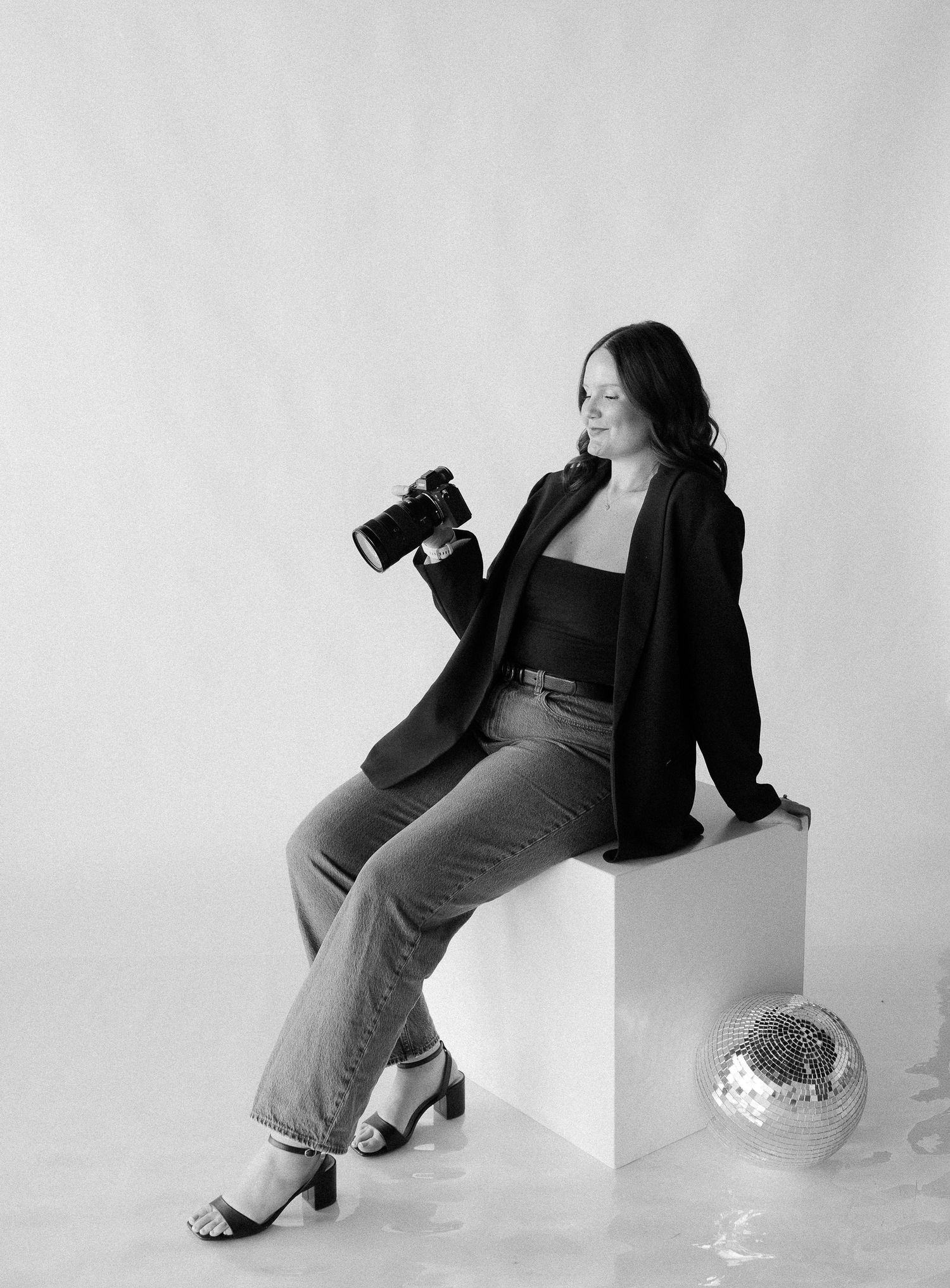 Professional photographer in casual attire poses with camera on white cube against plain background.