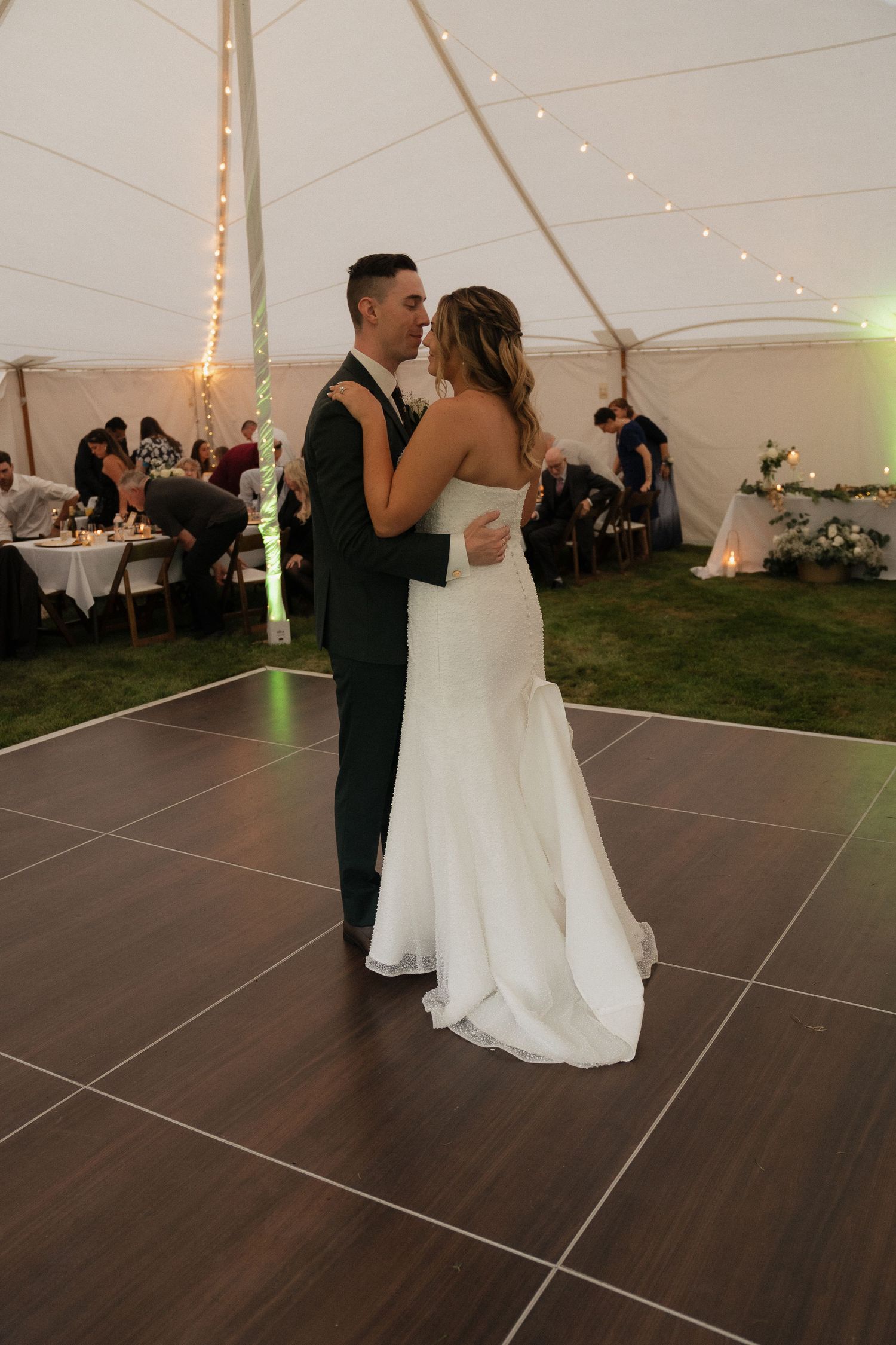 A couple shares their first dance in a tent reception with string lights and wooden dance floor.