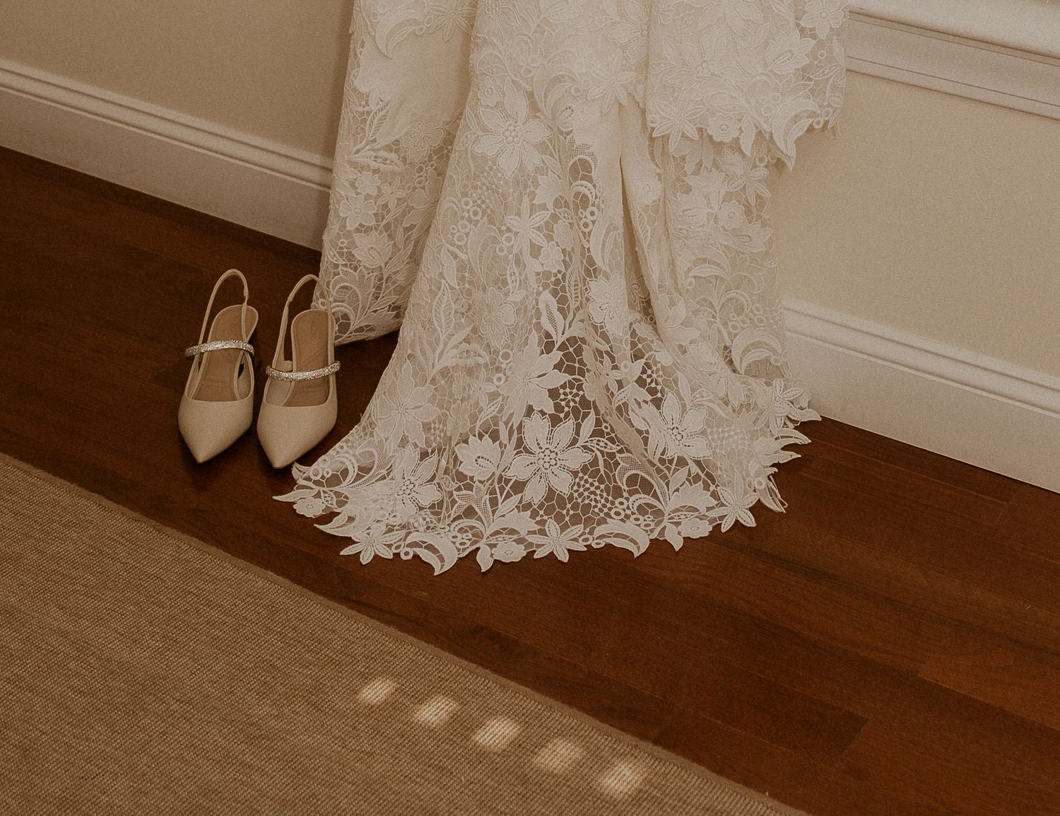 Elegant white lace wedding dress hem and beige bridal shoes on dark hardwood floor with sunlight streaming through.