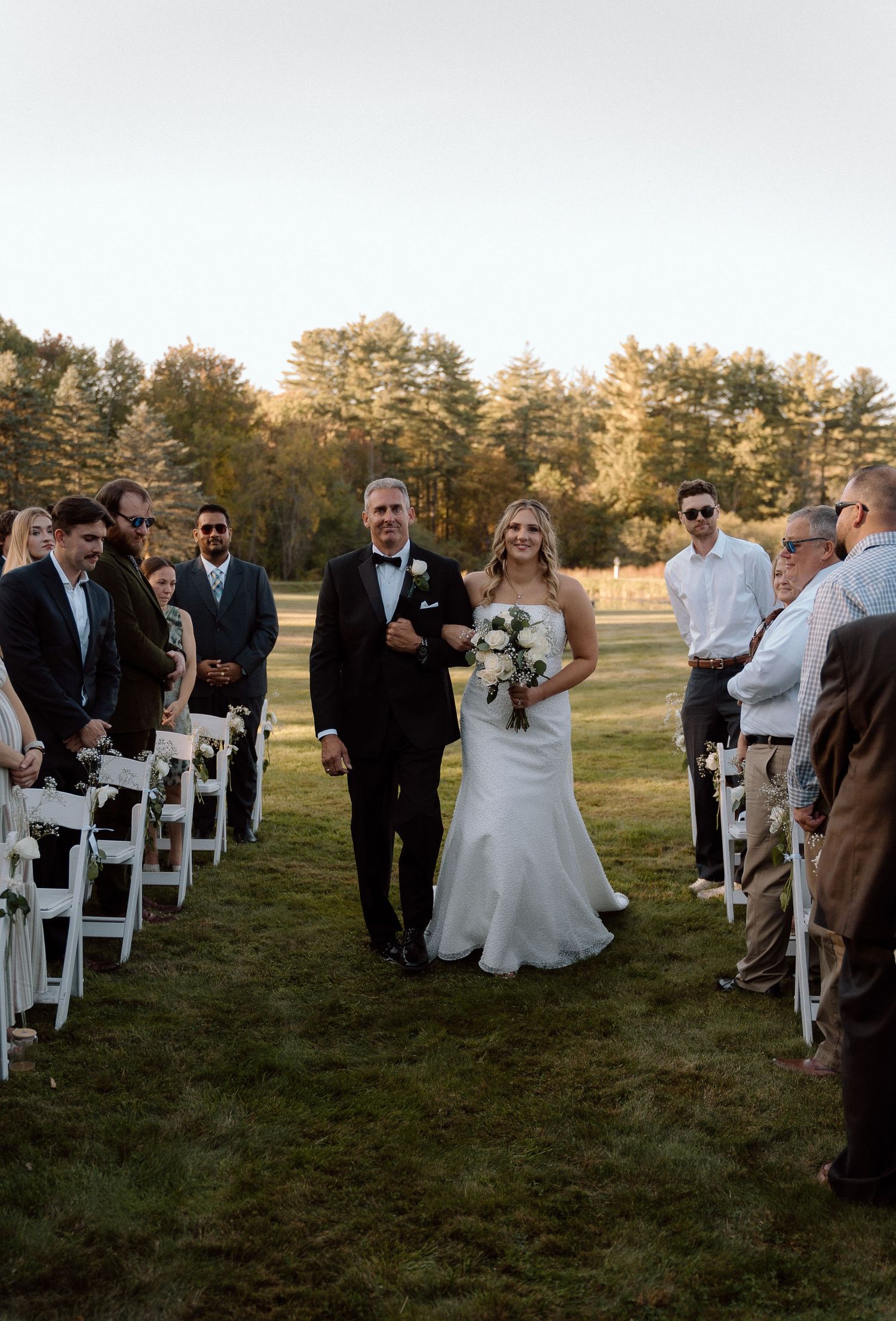 Wedding party and guests mingle on a lawn lined with white folding chairs during sunset.