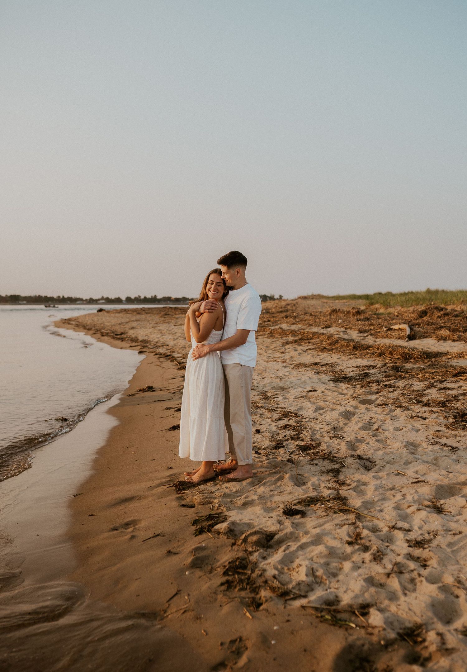 Romantic couple embracing on a sandy beach at sunset with calm waters in the background.