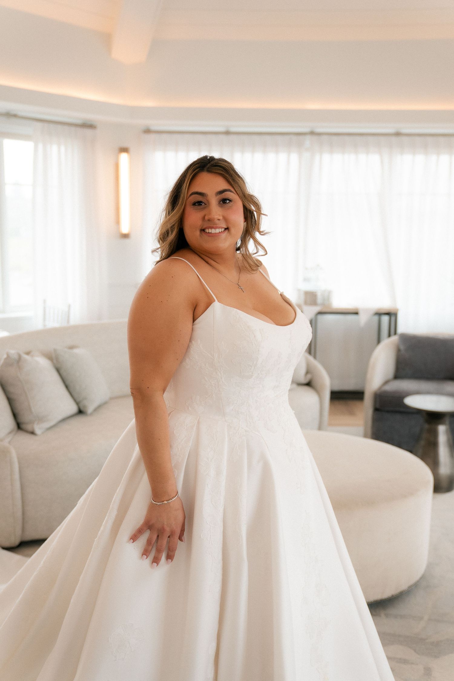 Smiling bride in elegant white spaghetti strap ball gown wedding dress in a bright hotel suite.
