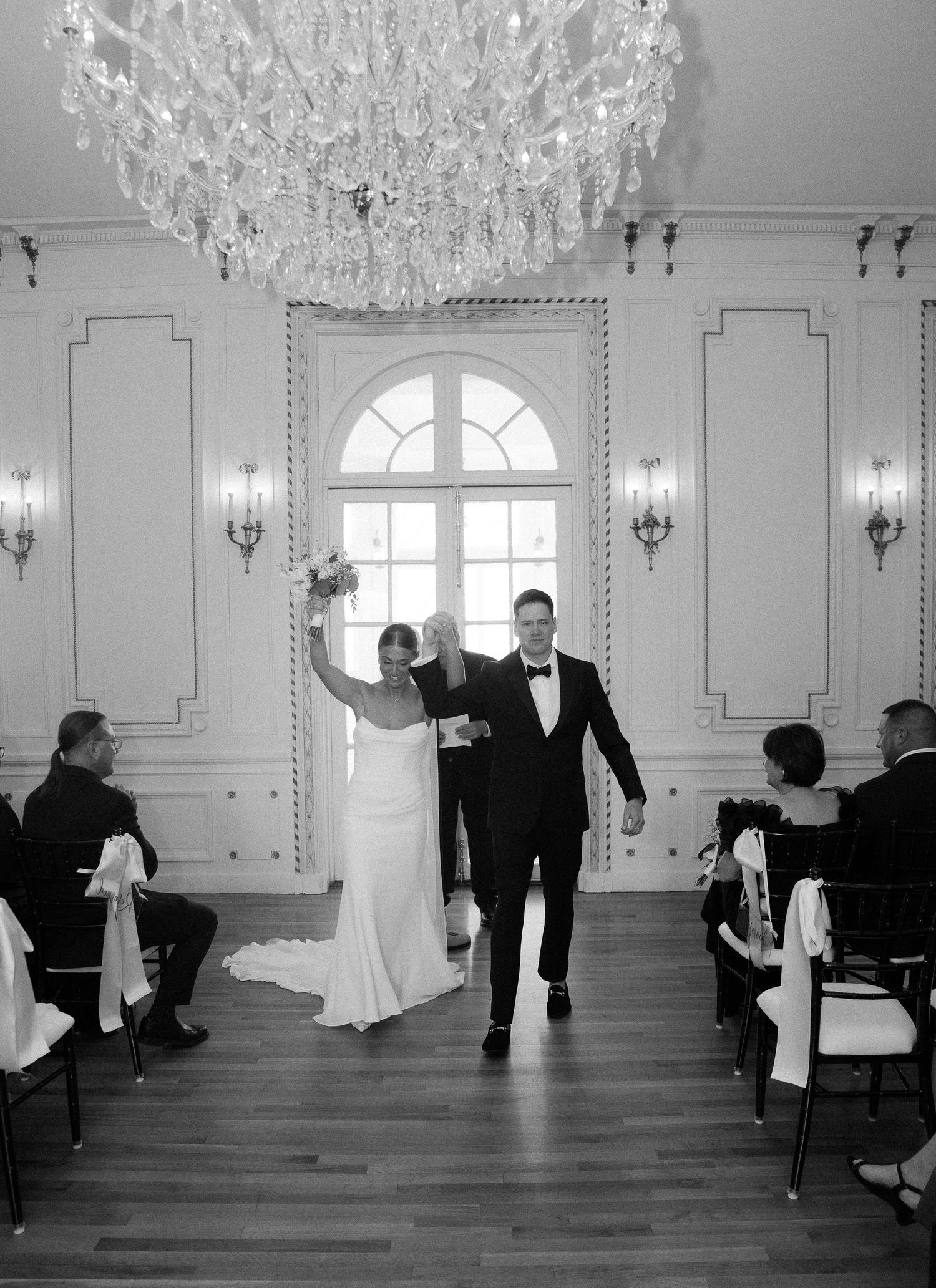 Bride and groom walk down the aisle in elegant ballroom with crystal chandelier after wedding ceremony.